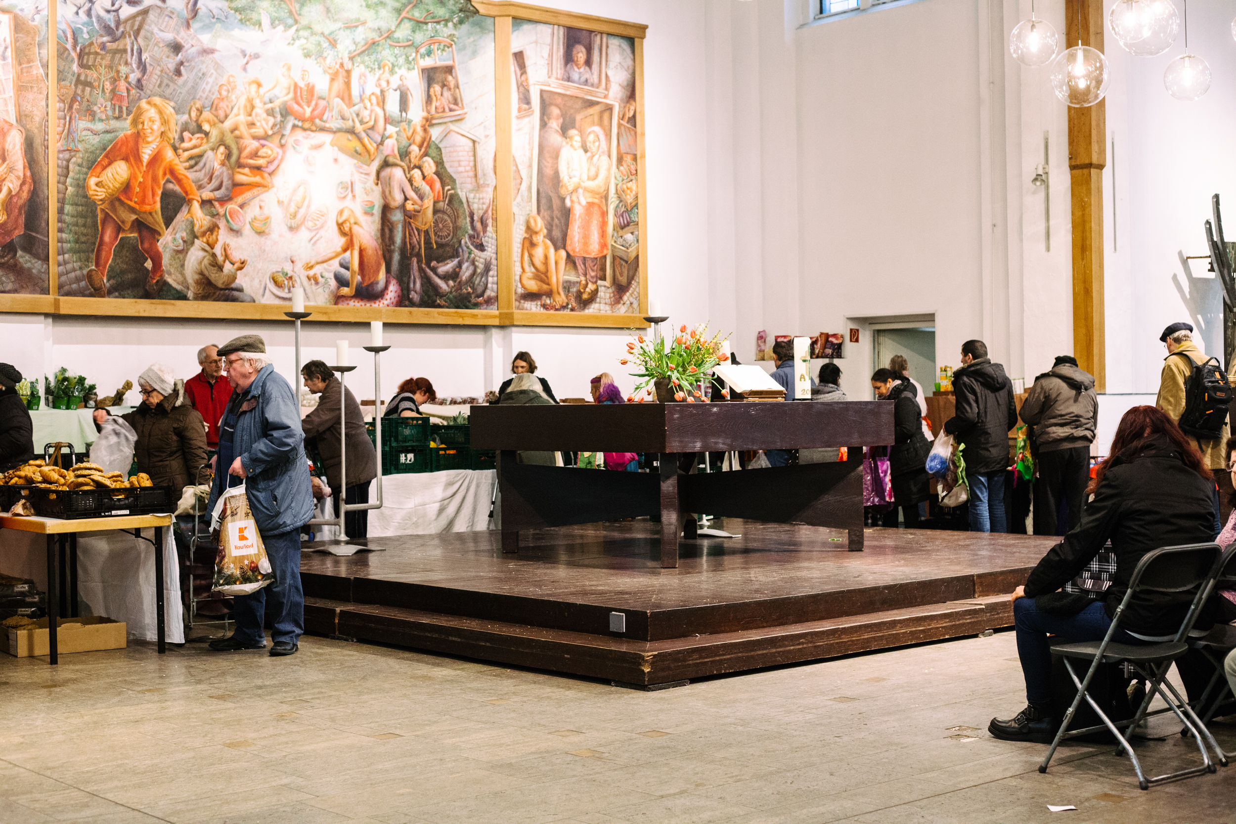 Mehrere alte Menschen stehen in einer Schlange rund um einen quadratischen Holzaltar und vor den Tischen mit dem Essen, darüber hängt ein großes Gemälde mit einer Picknickszene