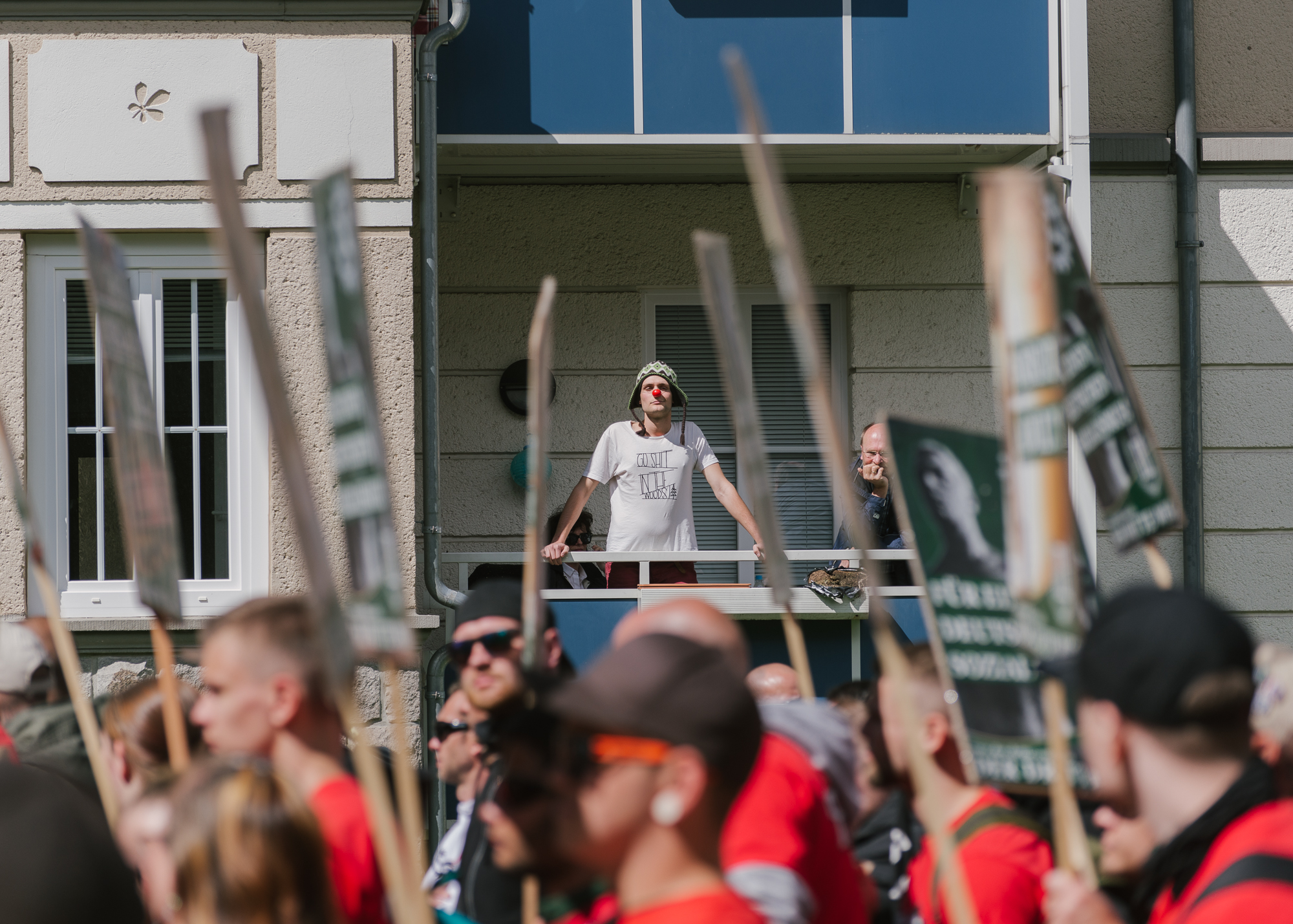 Junger Mann steht mit Clownsnase auf Balkon, im Vordergrund läuft die Demo des III. Wegs