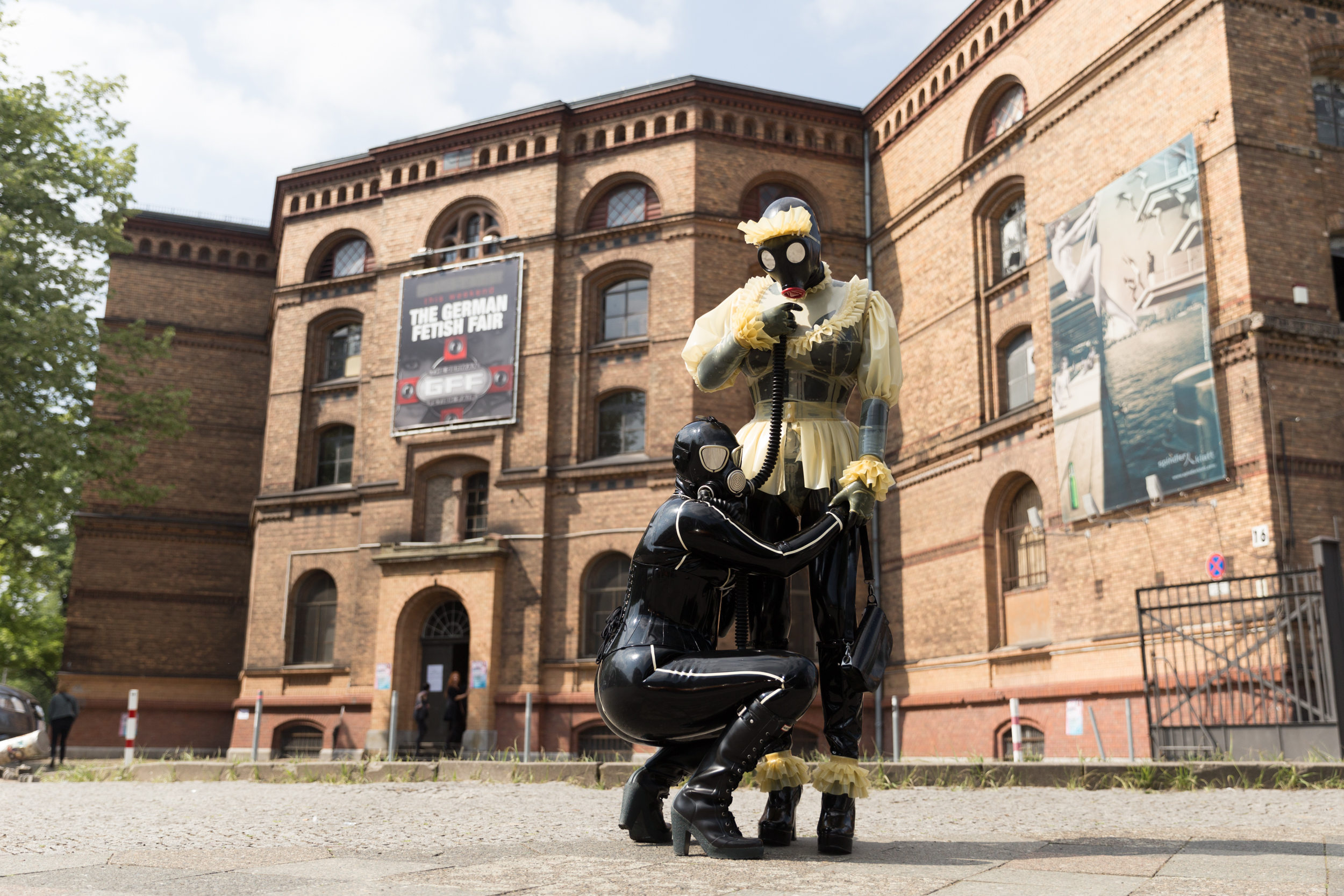 Eine Person in einem Ganzkörperlatexanzug kniet vor der stehenden Gummipuppe, die nach dem Schlauch der Gasmaske greift