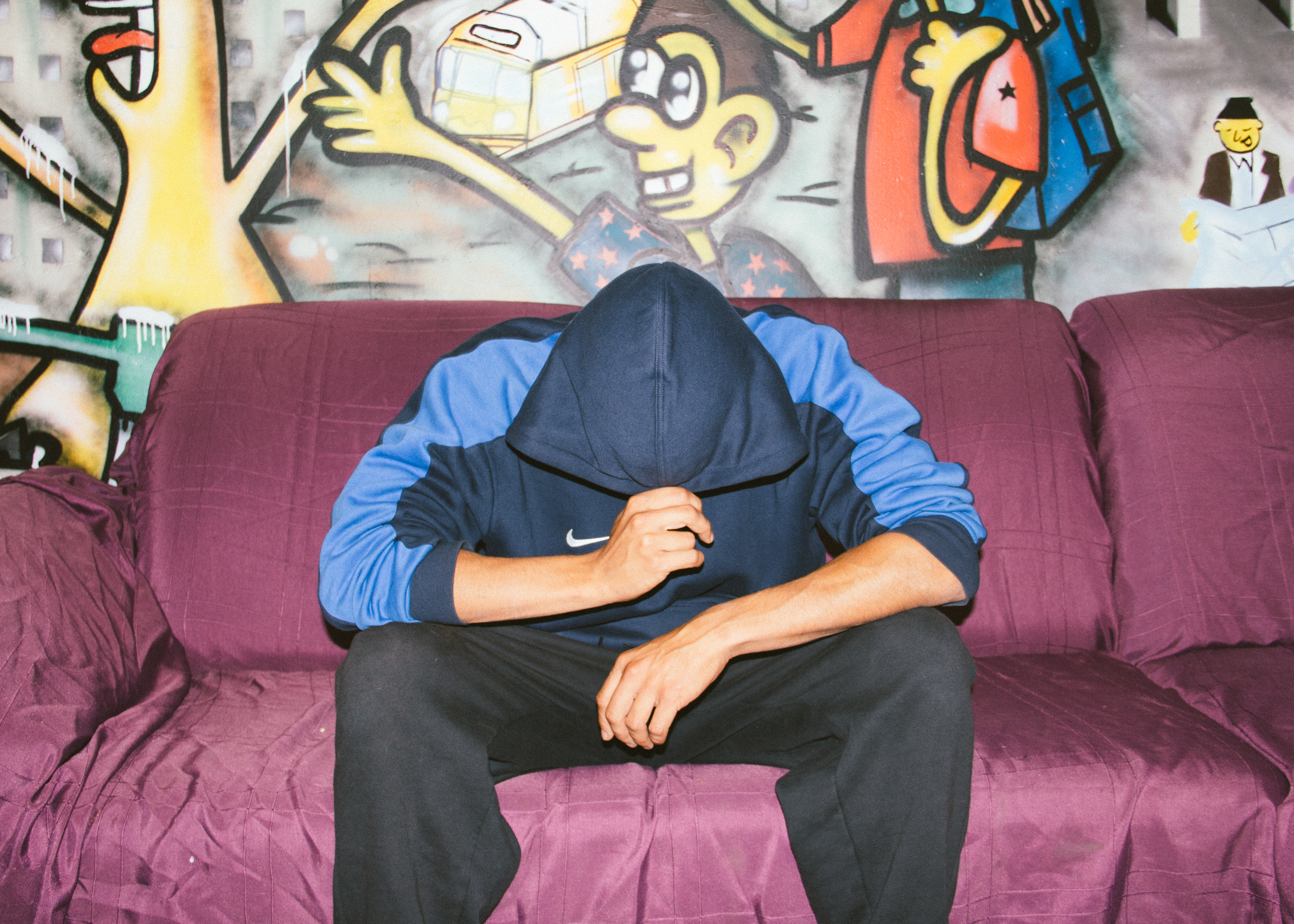 Ein Mann sitzt auf einer Couch vor einer mit Graffiti besprühten Wand und zieht die Kapuze seiner Trainingsjacke vors Gesicht