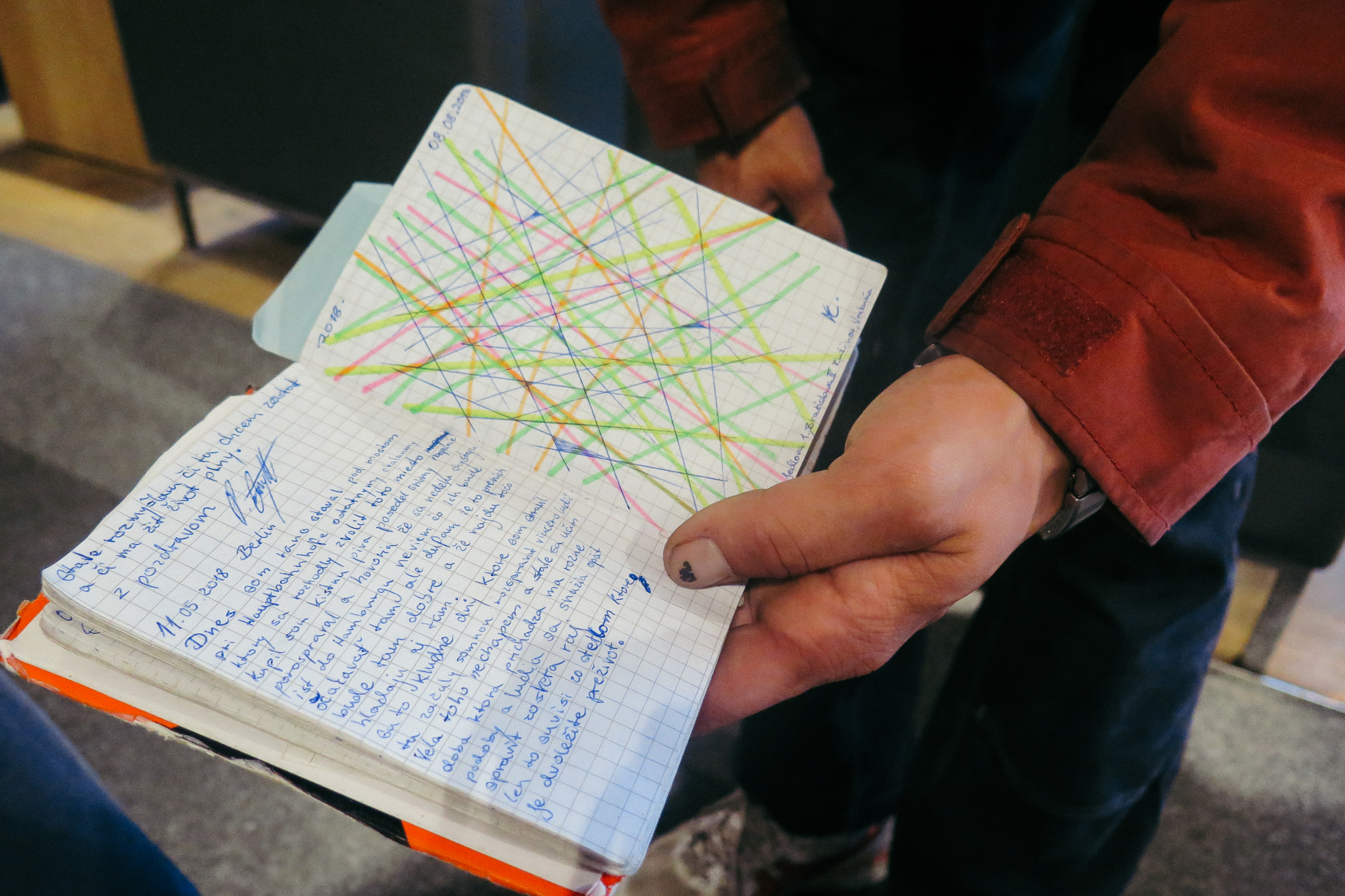 Eine Hand hält ein kleines Buch mit kariertem Papier hoch, auf der linken Seite ist ein Tagebucheintrag notiert, auf der rechten Seite eine abstrakte Zeichnung bunter Linien