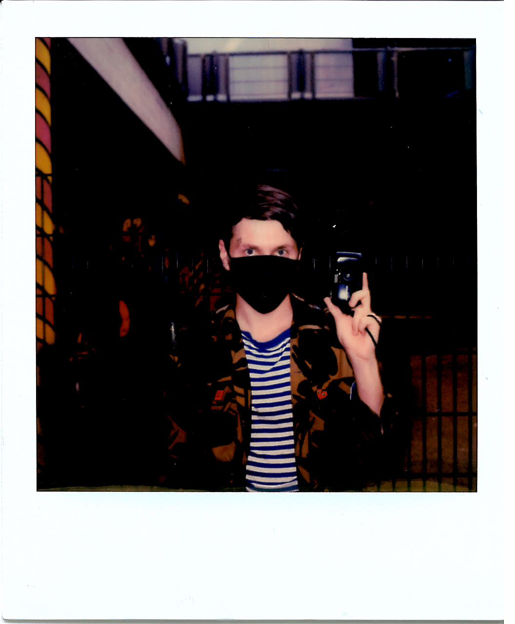 Ein Polaroid auf dem ein junger Mann mit Maske vor Mund und Nase eine Analogkamera hochhält