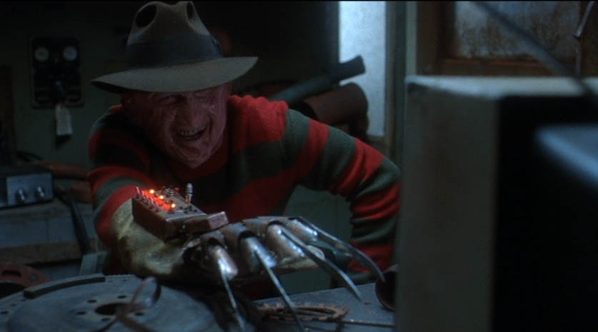 a smiling Freddy Krueger leans forward over a desk toward a TV, his knife glove augmented with an electronic device
