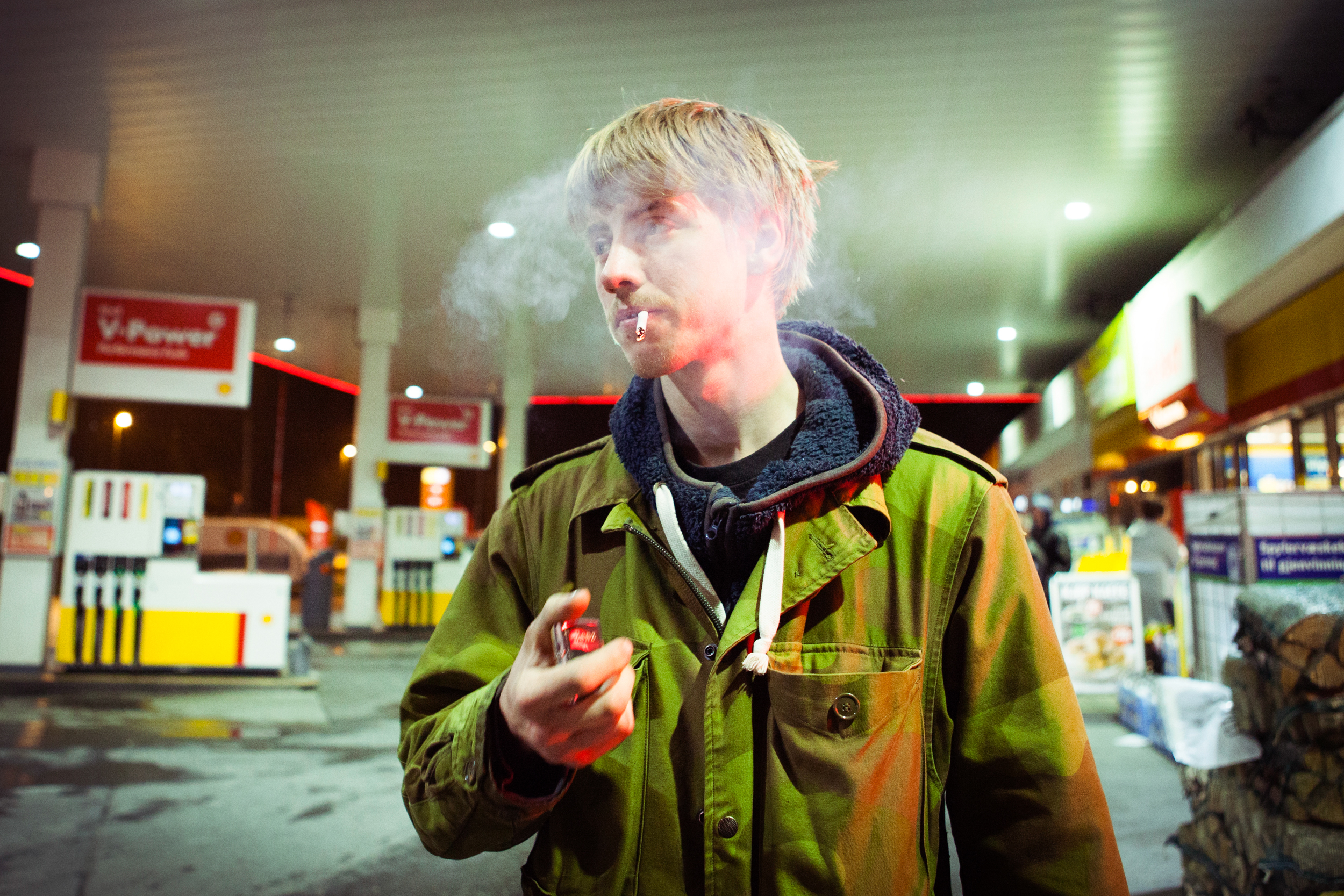 En ung mand på en tankstation. Han har lyst hår, er iført armygrøn jakke og ryger en cigaret
