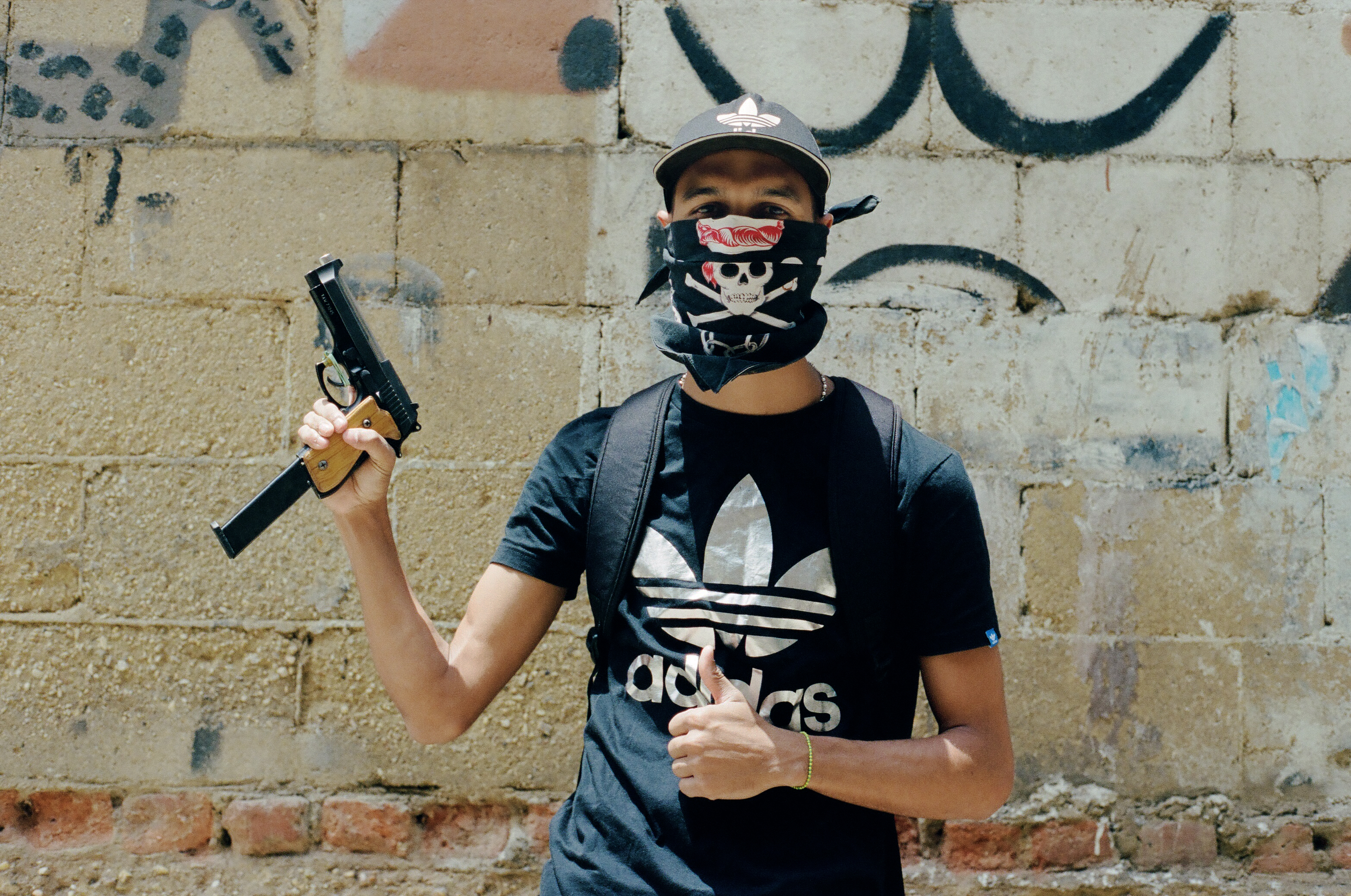 Ein Mann mit Basecap, T-Shirt und einem Tuch vor dem Gesicht hält eine Pistole hoch und reckt den Daumen der anderen Hand