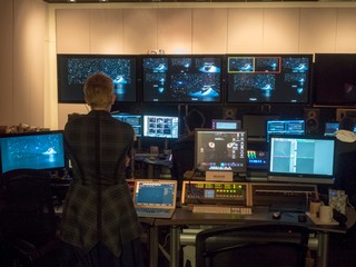 Rehearsal image, Producer Lisa Niedermeyer stands with her back to us and facing a dozen monitors in the video control room of EMPAC rehearsing the livestream of DESCENT. Five different cameras show the opening image of Andromeda among the stars from different points of view. The angle framed is red is the one streaming, and angle framed is yellow is cued up to go next.