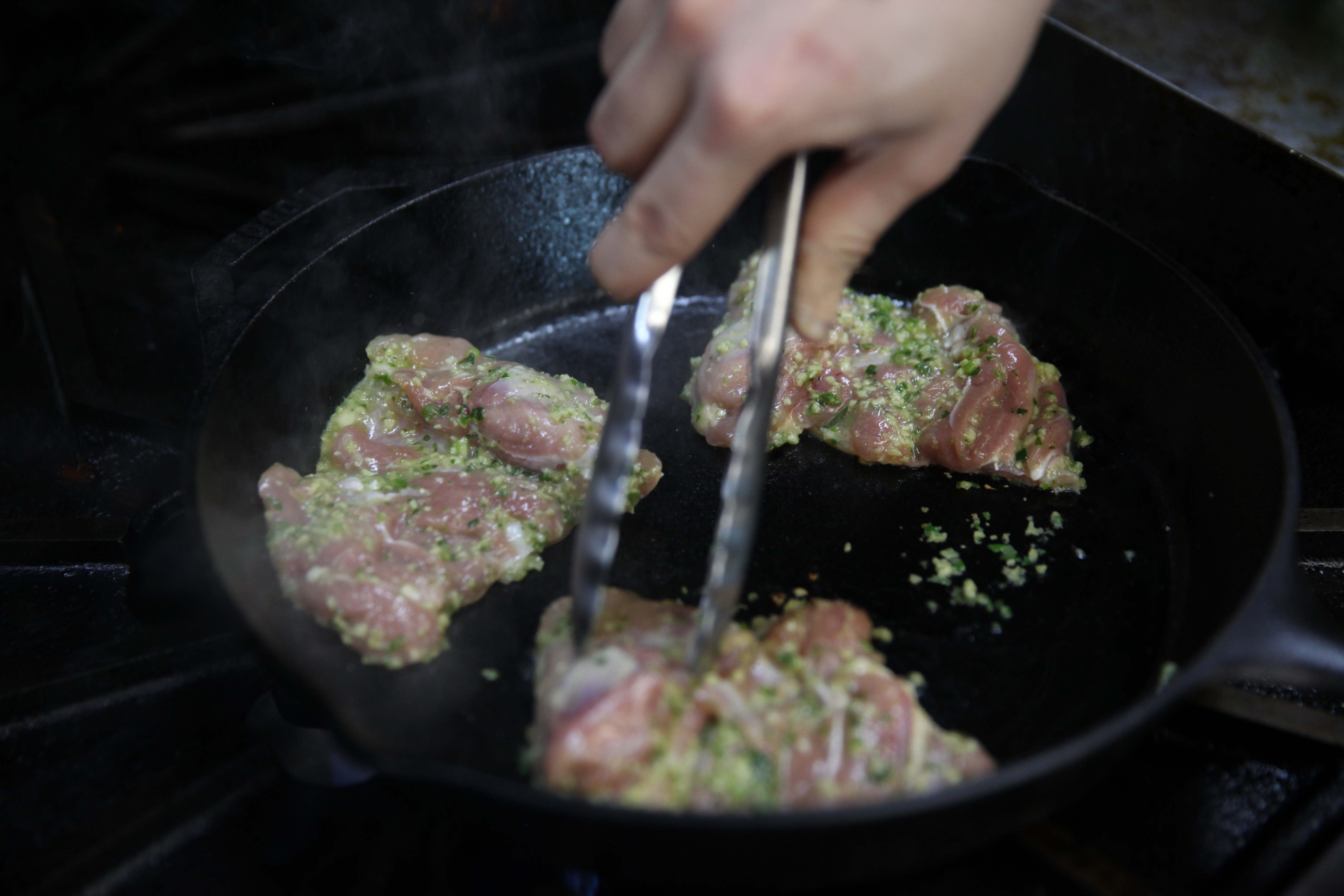 chicken thighs searing in a hot pan and a hand pushing them down with tongs