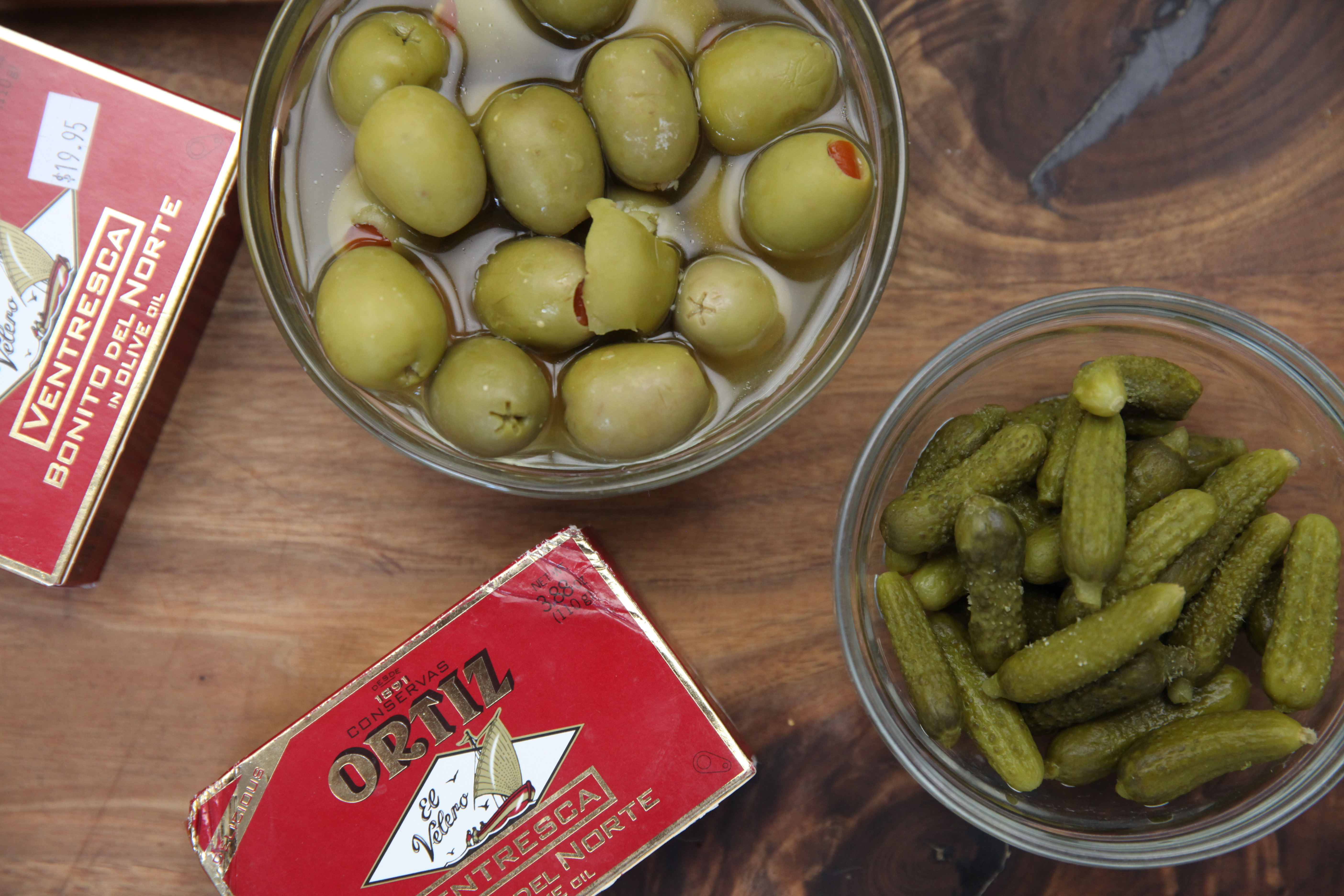 cans of ortiz ventresca tuna, a bowl of green olives with pimento, and a bowl of cornichons