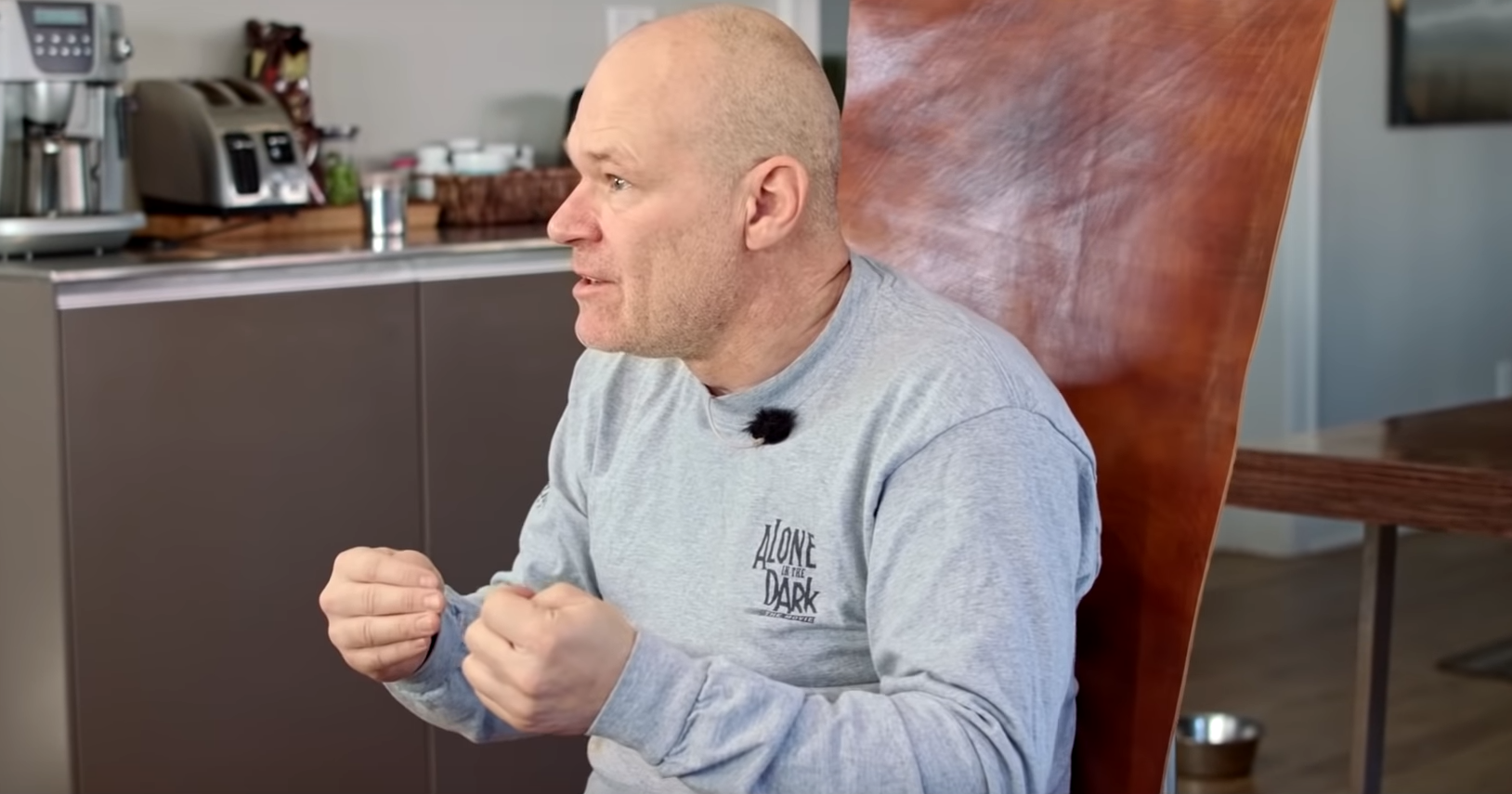 Uwe Boll looking cheerful and slightly punchy while wearing a pale gray Alone in the Dark sweater and explaining something to a figure off camera.