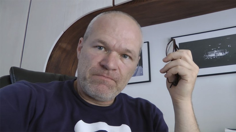 Uwe Boll looking supercilious while holding a folded pair of glasses and glaring down at the camera.