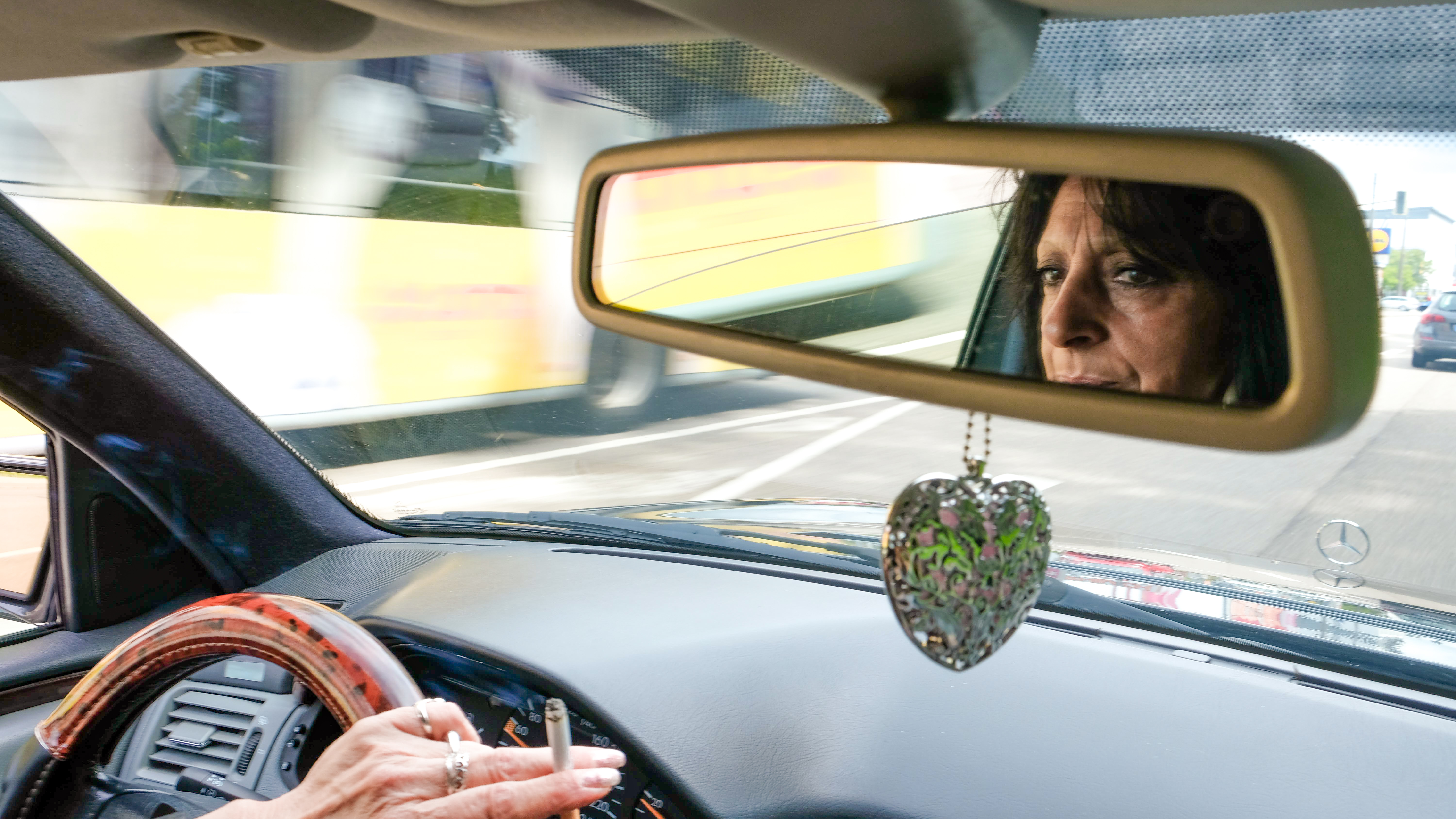 Taxifahrerin Eva Leidinger fährt ihr Taxi. Sie spiegelt sich im Rückspiegel, an dem ein Schmuckherz aus Metall baumelt. In ihrer Hand brennt eine Zigarette. Sie raucht im Auto.