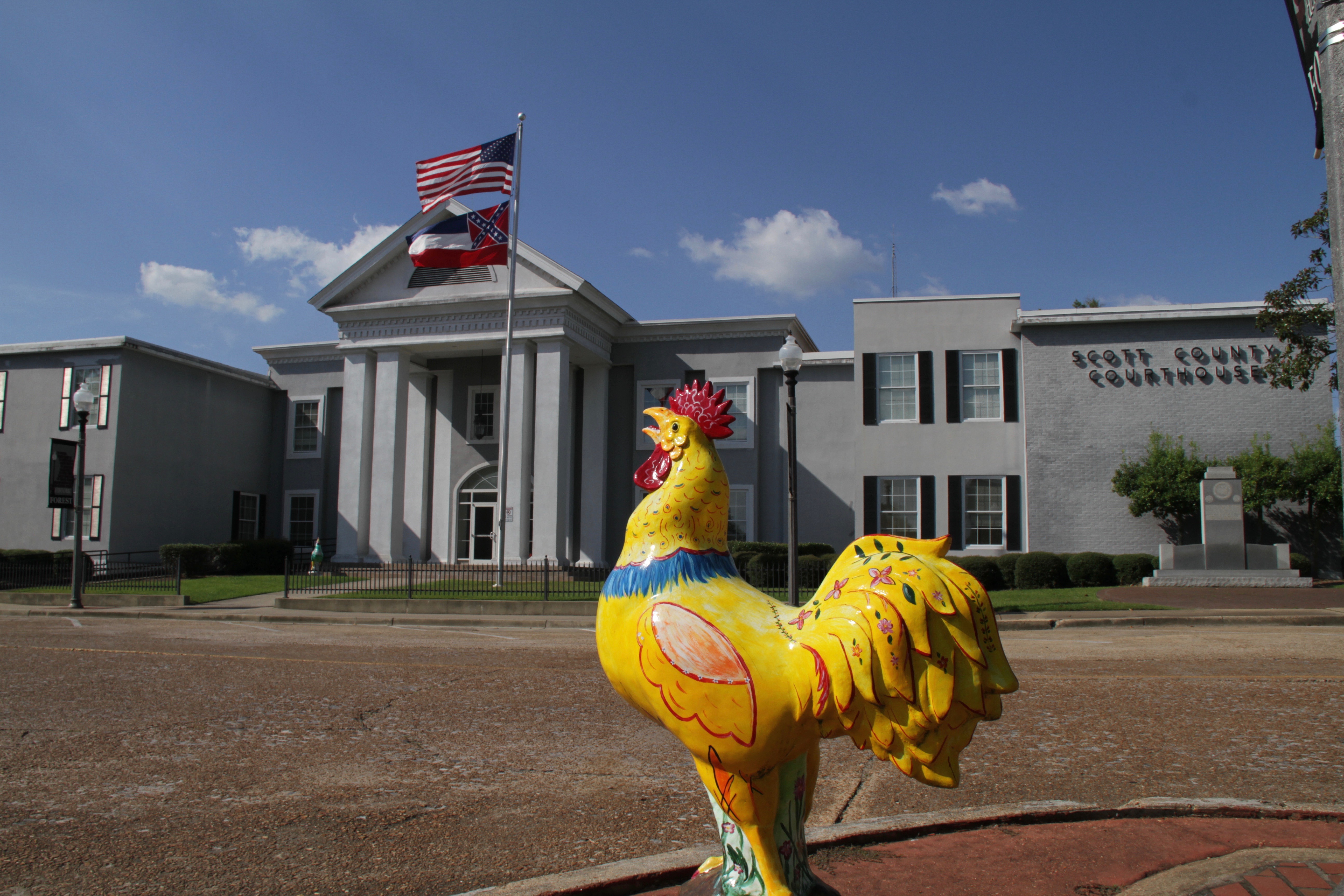 A chicken statue in front of a courthouse flying the Mississippi state flag.
