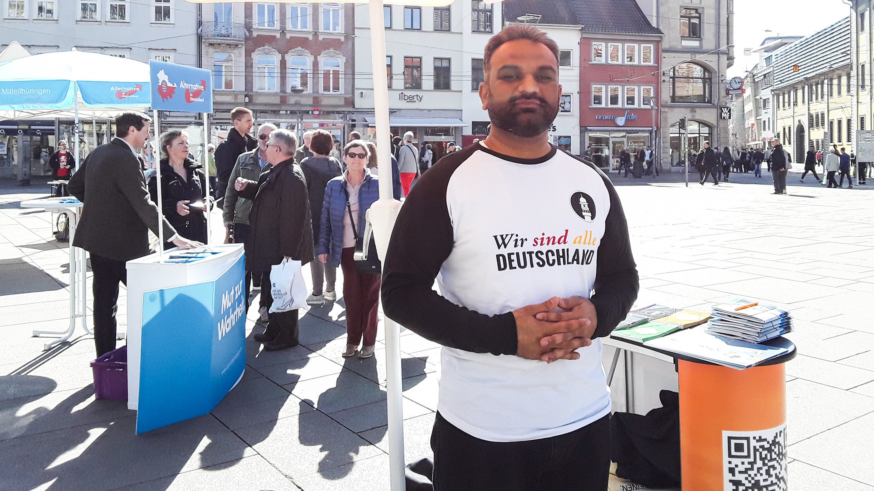 Ein dunkelhäutiger Mann steht vor einem Stand an einem Marktplatz, auf seinem T-Shirt steht: Wir sind alle Deutschland. Hinter ihm sieht man einen AfD Stand