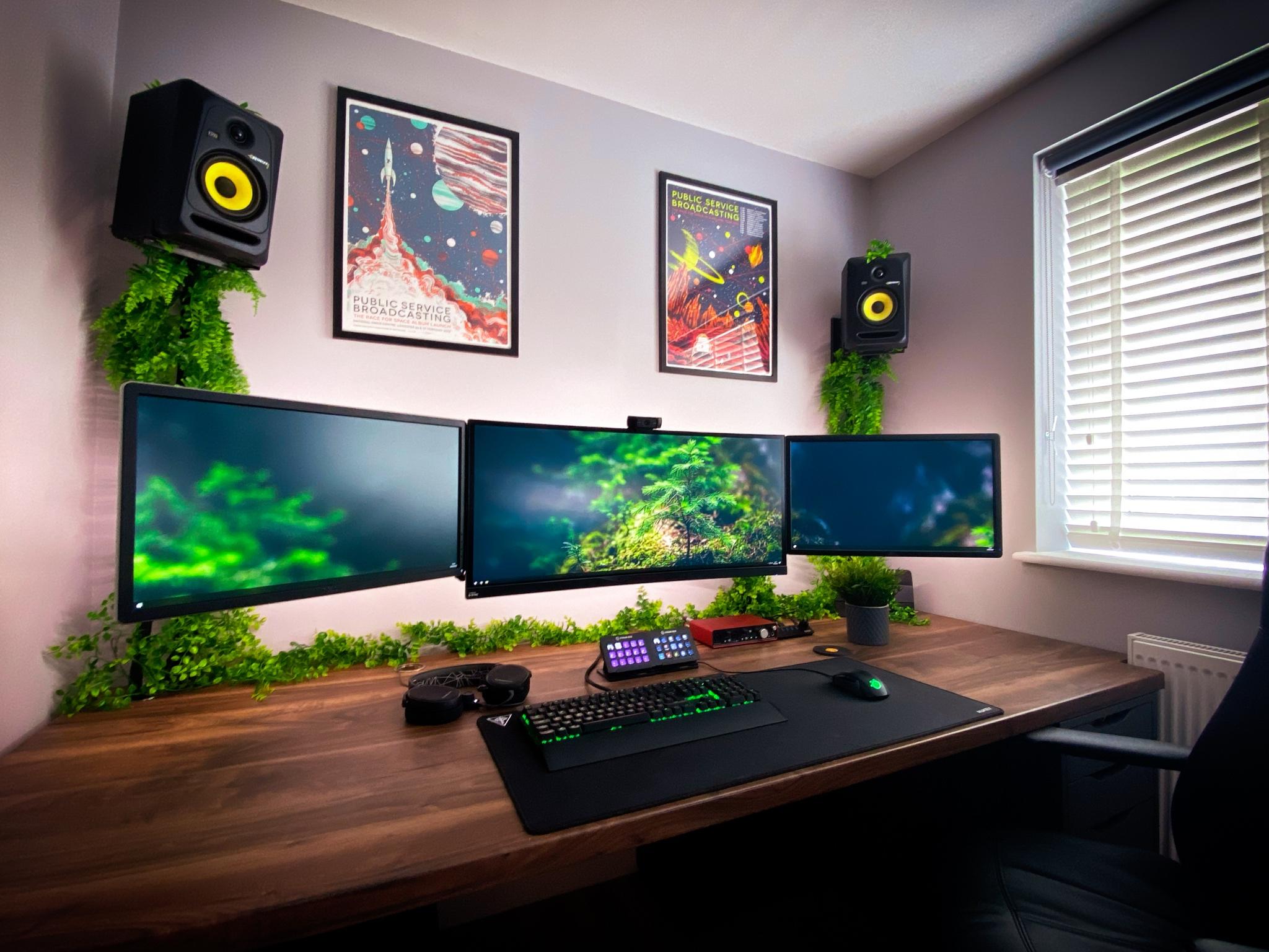 A three-monitor display encompasses almost the width of a large, room-spanning desk. Ceiling-mounted speakers dangle above, framing both the listener and a pair of sci-fi posters.