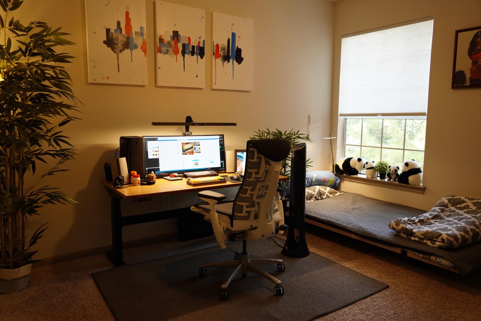 A cozy bedroom office with stuffed pandas framing the window above a low cot next to a warmly lit, uncluttered desktop with a Herman Miller office chair