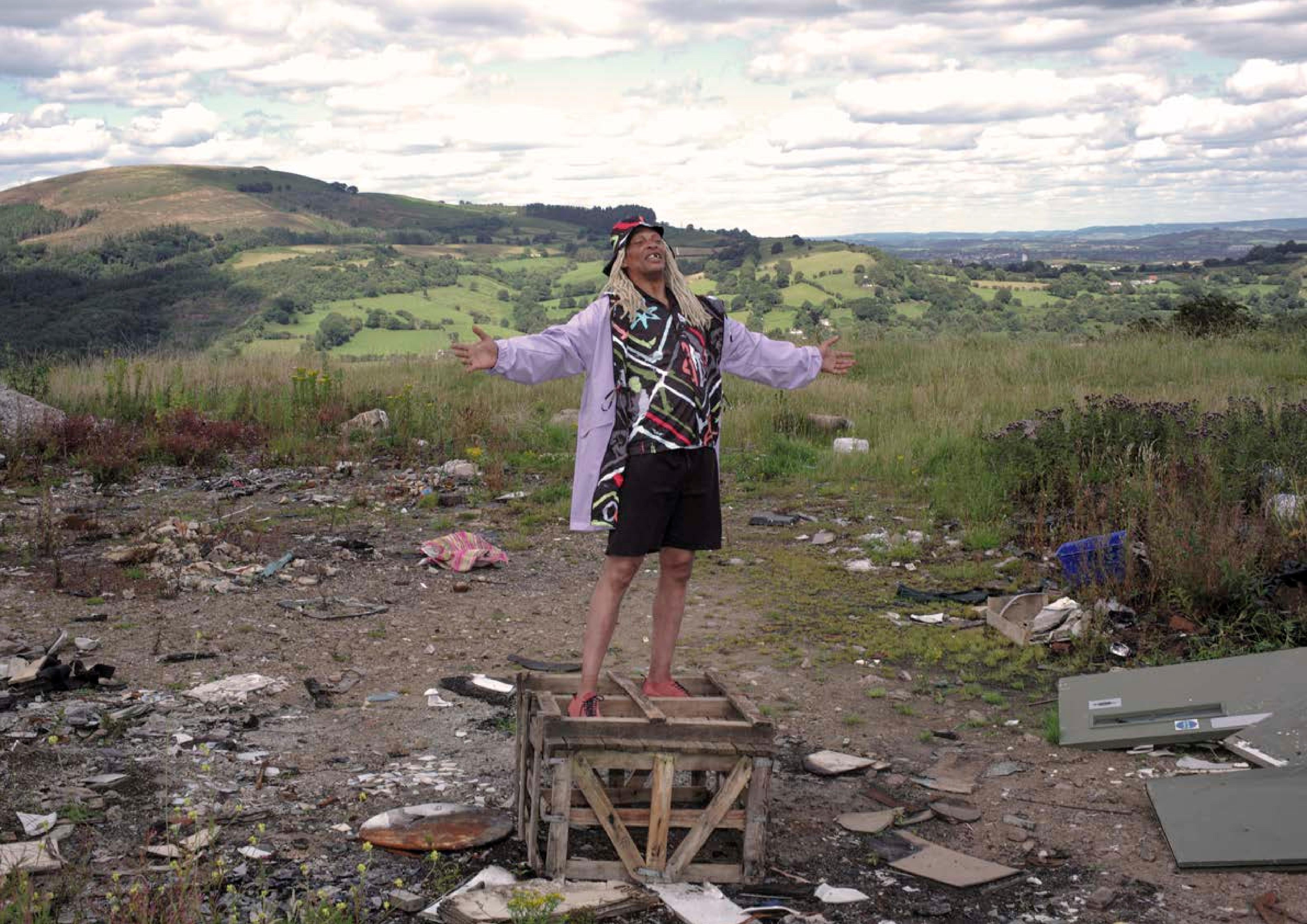 A man standing on a wooden crate in colourful clothes in a heavily littered landscape