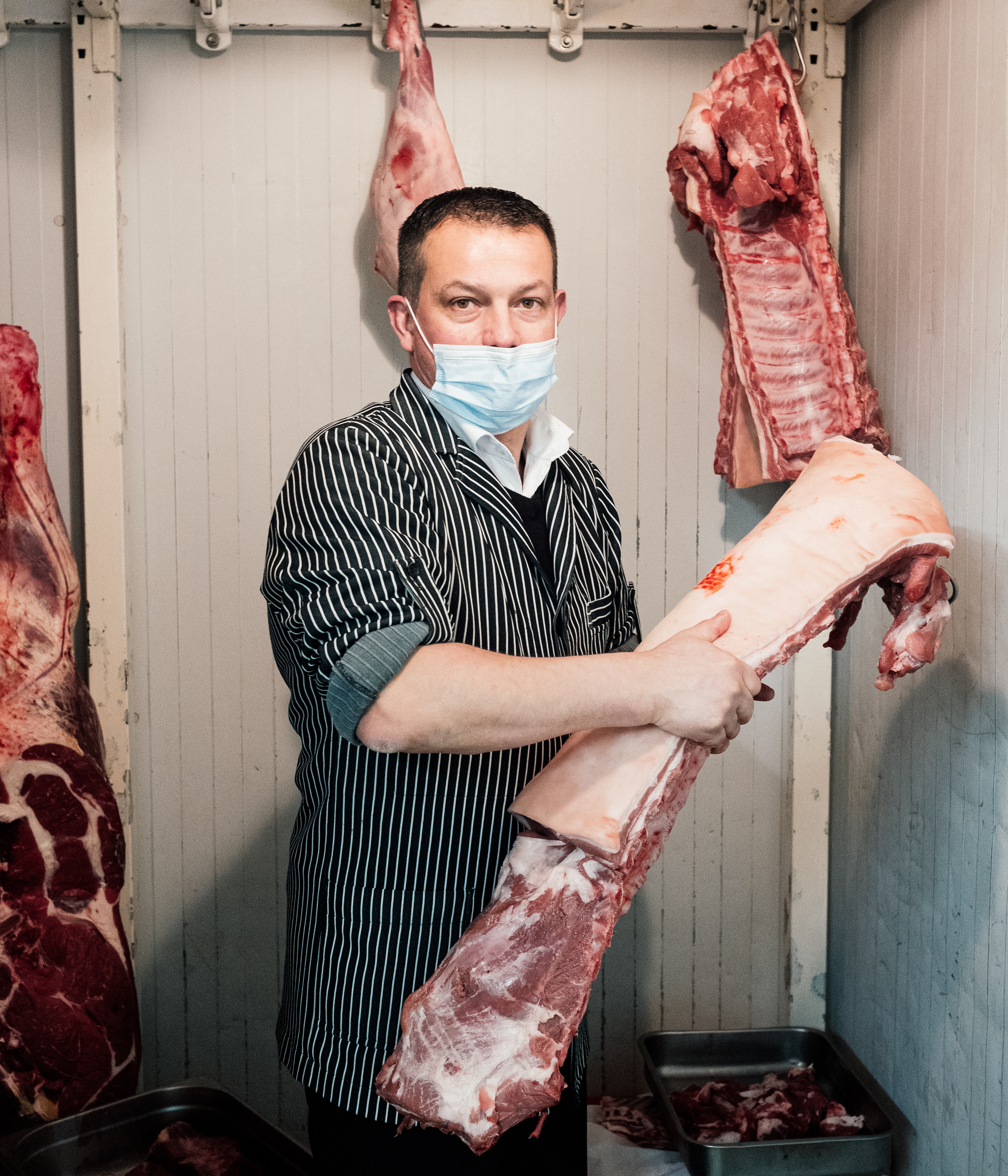 Sébastien Freteau wearing a black and white pinstripe shirt and a mask, handling a large piece of pork meat.