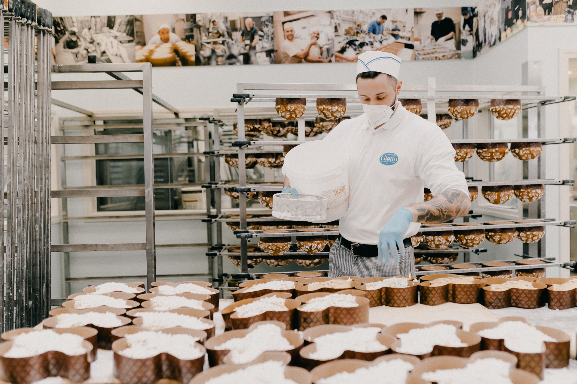 Giotto Bakery – A man wearing gloves between the racks with a large container and sprinkling the colombe.
