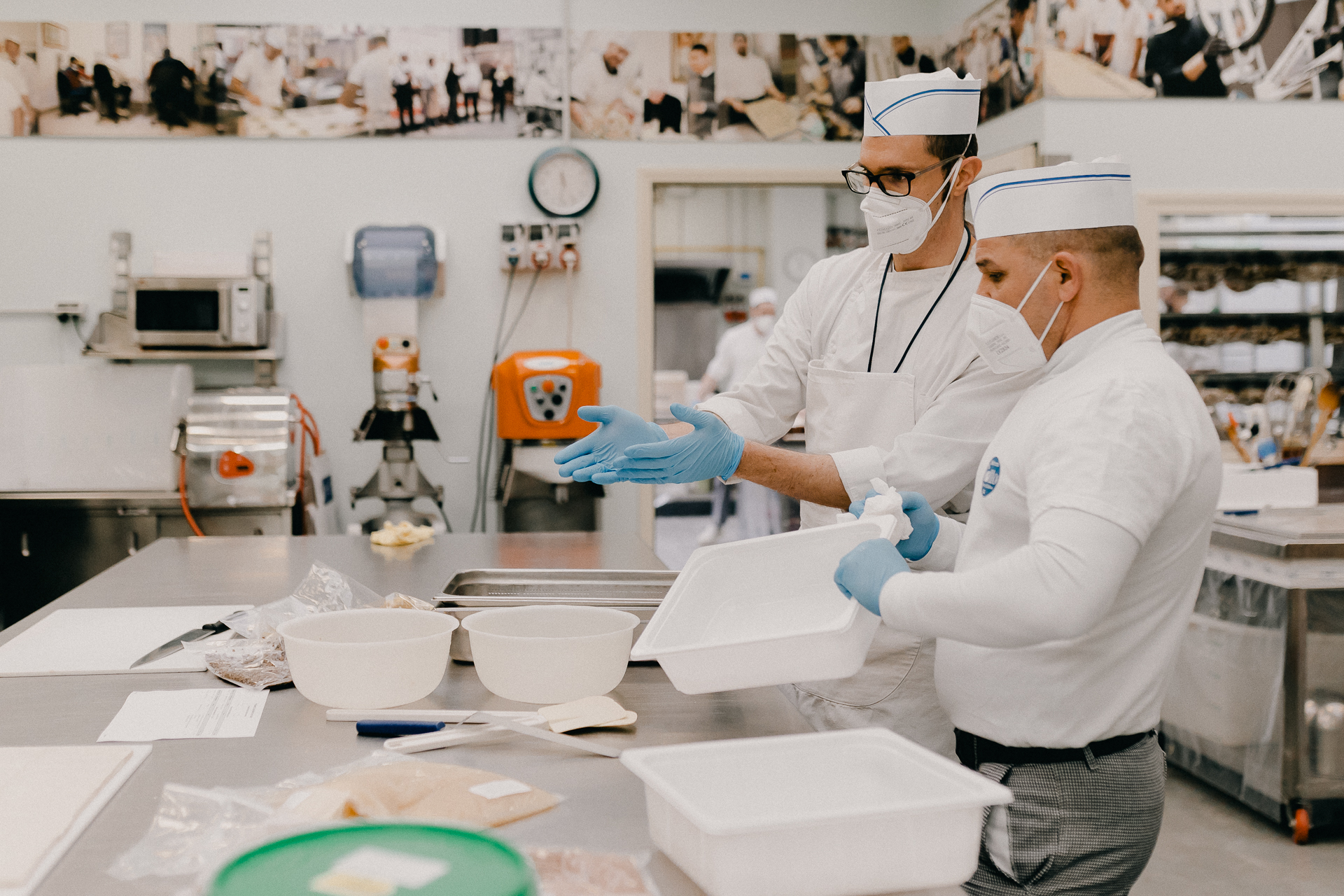 Giotto Bakery – One man talking to another, gesturing towards some plastic bowls on a work table as the other holds a plastic tray.