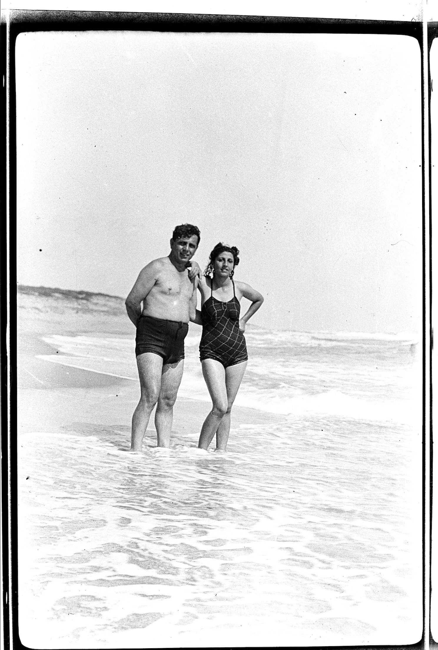 Kegham Djeghalian, Gaza photographer – couple in vintage bathing suits hugging and posing for a photo with their feet in the sea.
