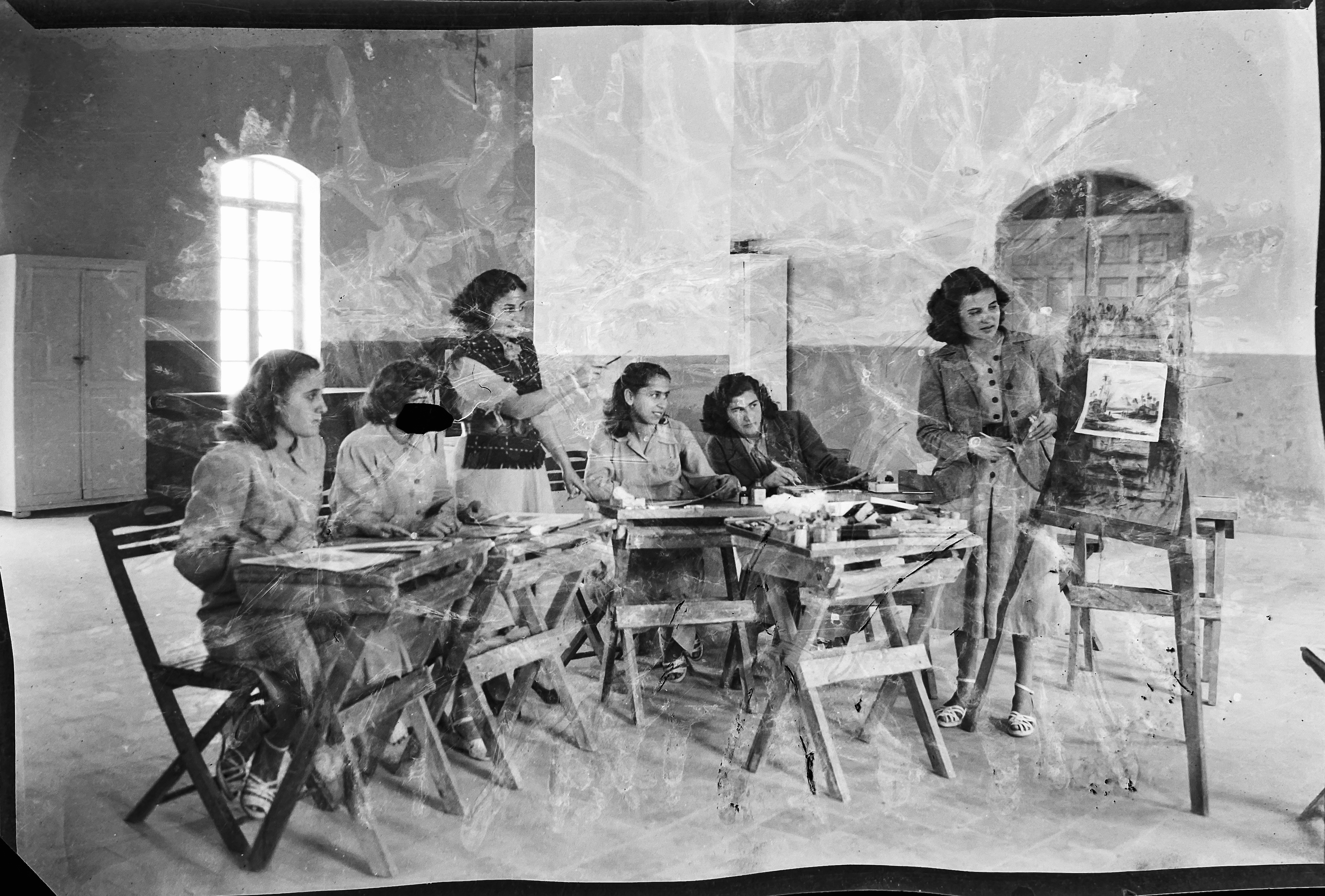 Kegham Djeghalian, Gaza photographer – six women sitting at desks in a large room, talking about a picture or a painting placed on an easel.