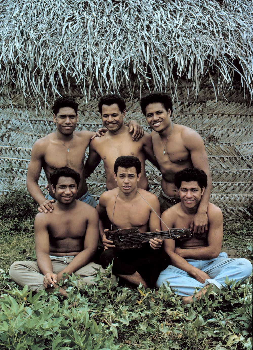 Keenam remaja duduk di depan gubuk. Seseorang memegang ukulele buatannya sendiri.