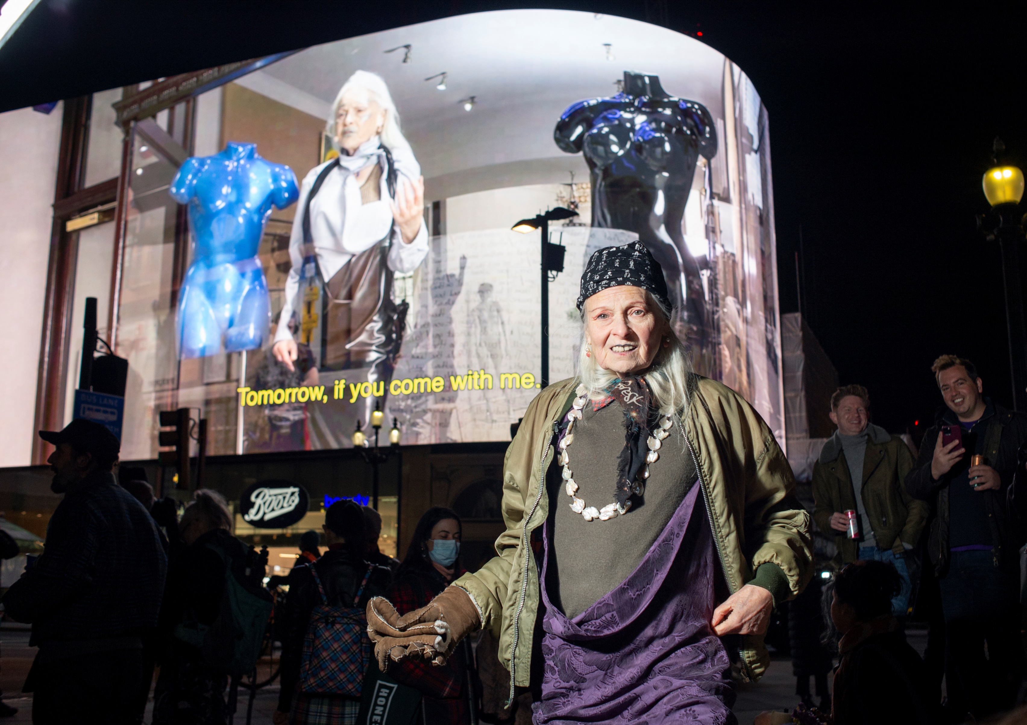 Vivienne Westwood stood in front of the Piccadilly Circus lights on her 80th birthday. Behind her, a video of her performing a reading plays. 