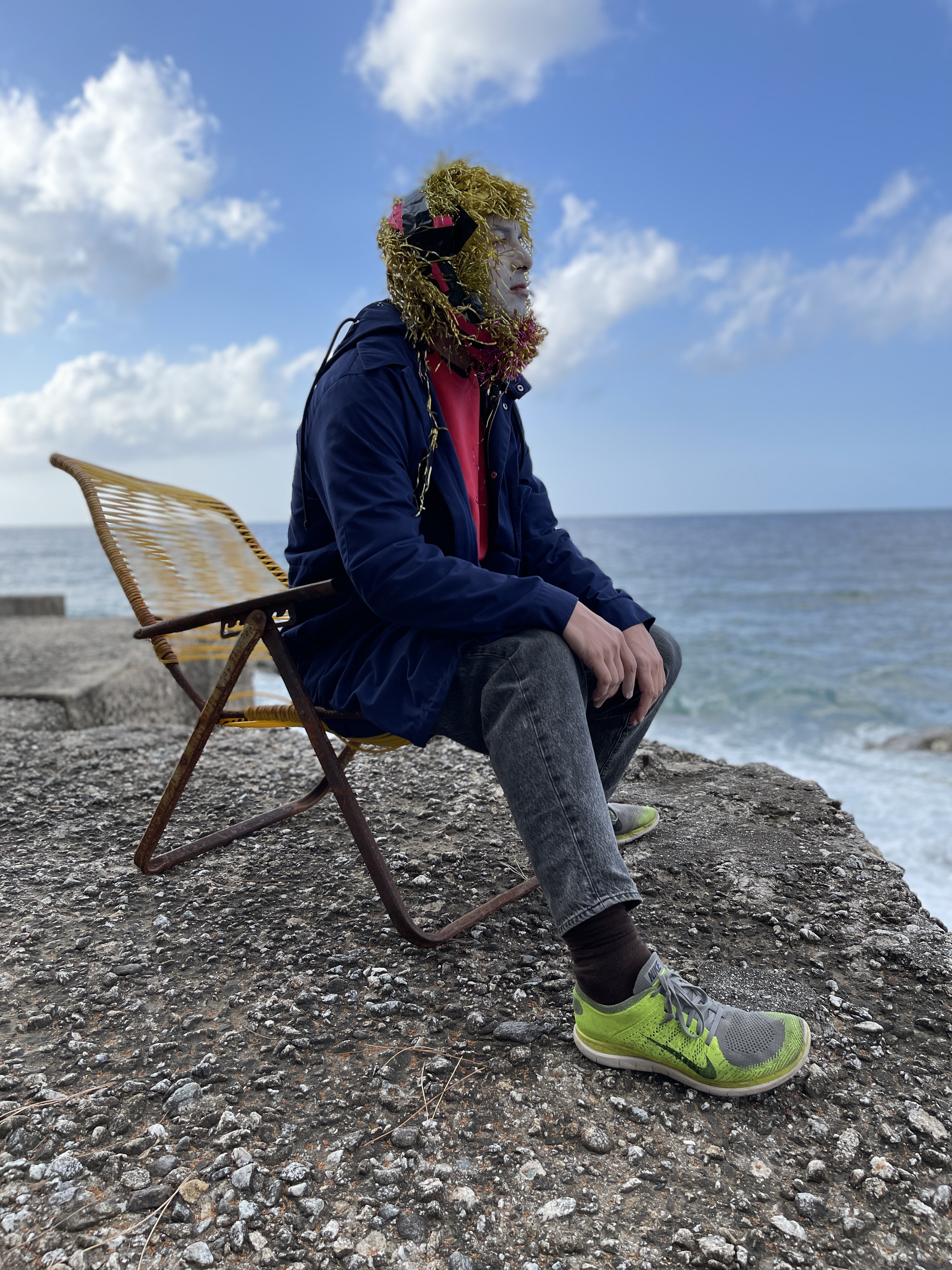 Nico vascellari seduto su una roccia con un copricapo di piume d'oro su una spiaggia di sassi