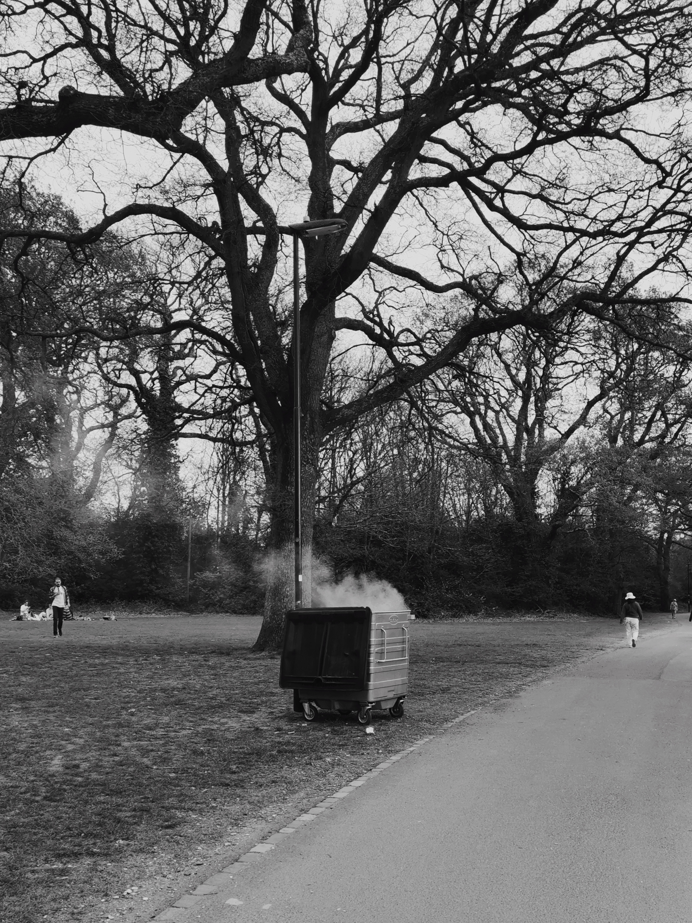 a large smoking bin in a public park, shot in black-and-white with an onlooker in the distance