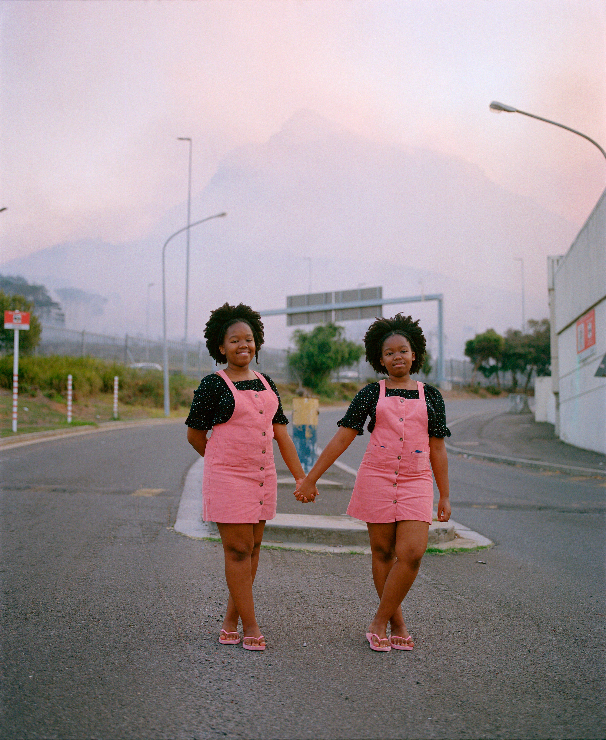 twin girls wearing the same pink pinafore dresses stand in the road in front of Table Mountain