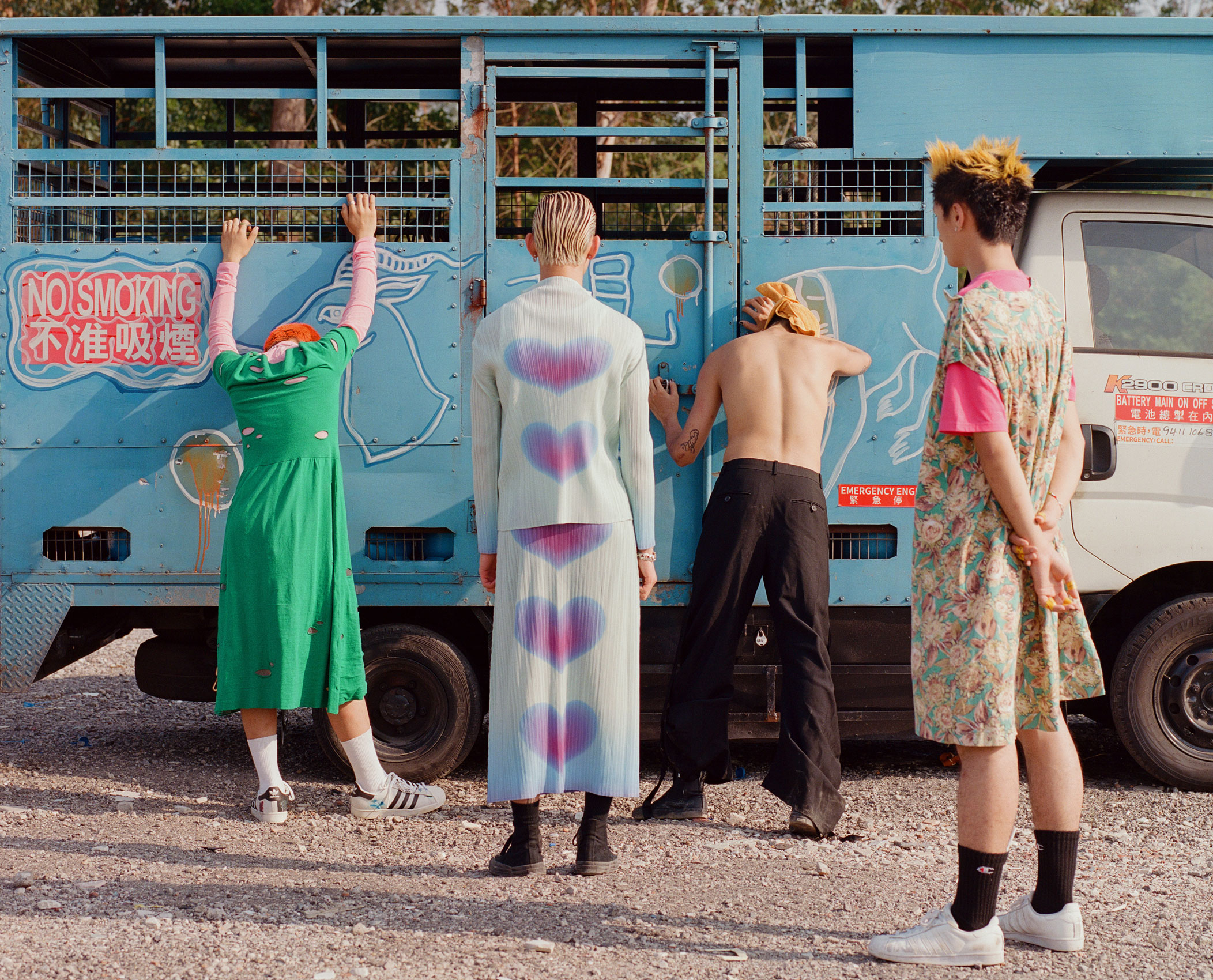 four boys wearing dresses stand with their backs to the camera, in front of an old pale blue truck