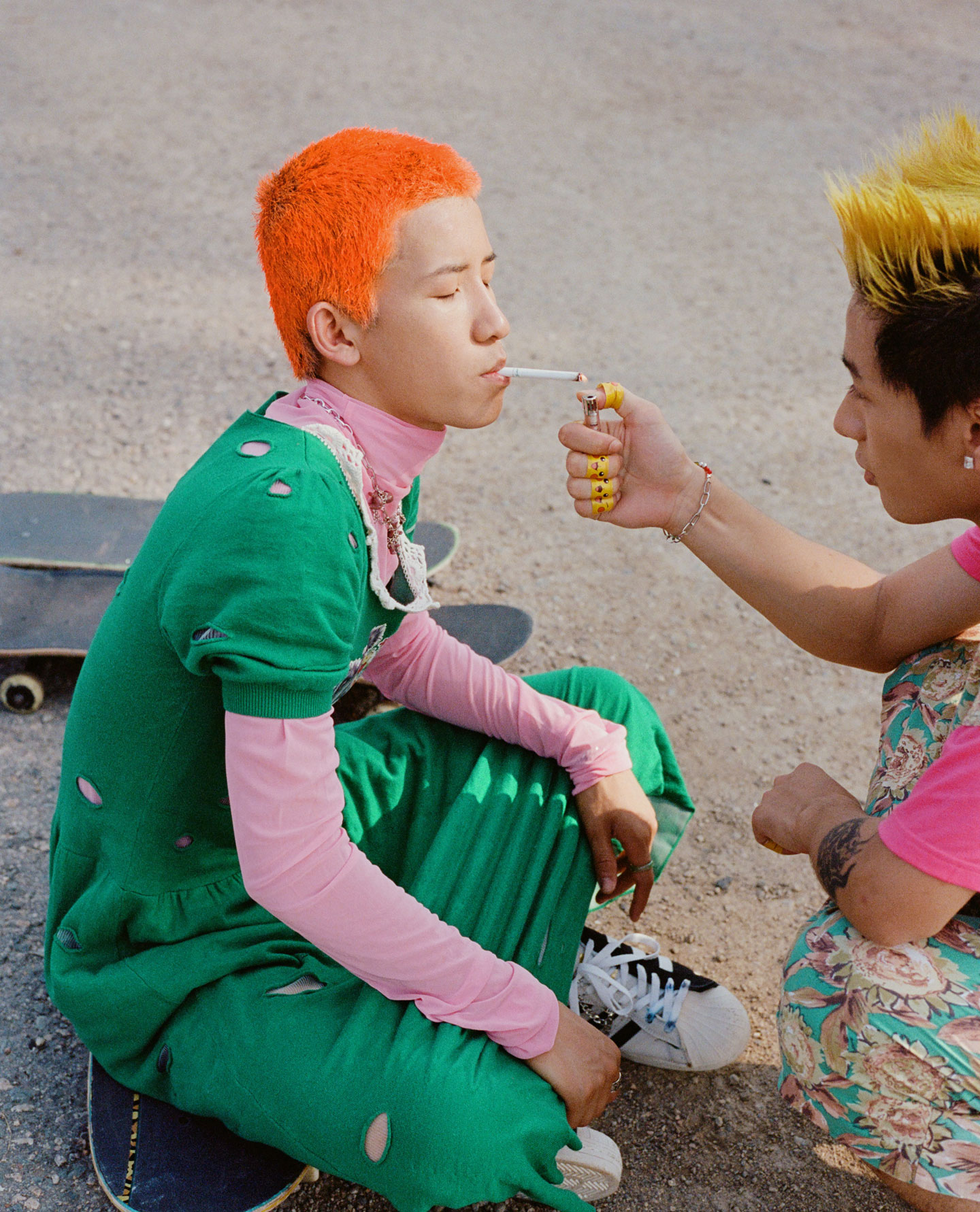 two young men with colourful hair sit on skateboards, one lights the other's cigarette