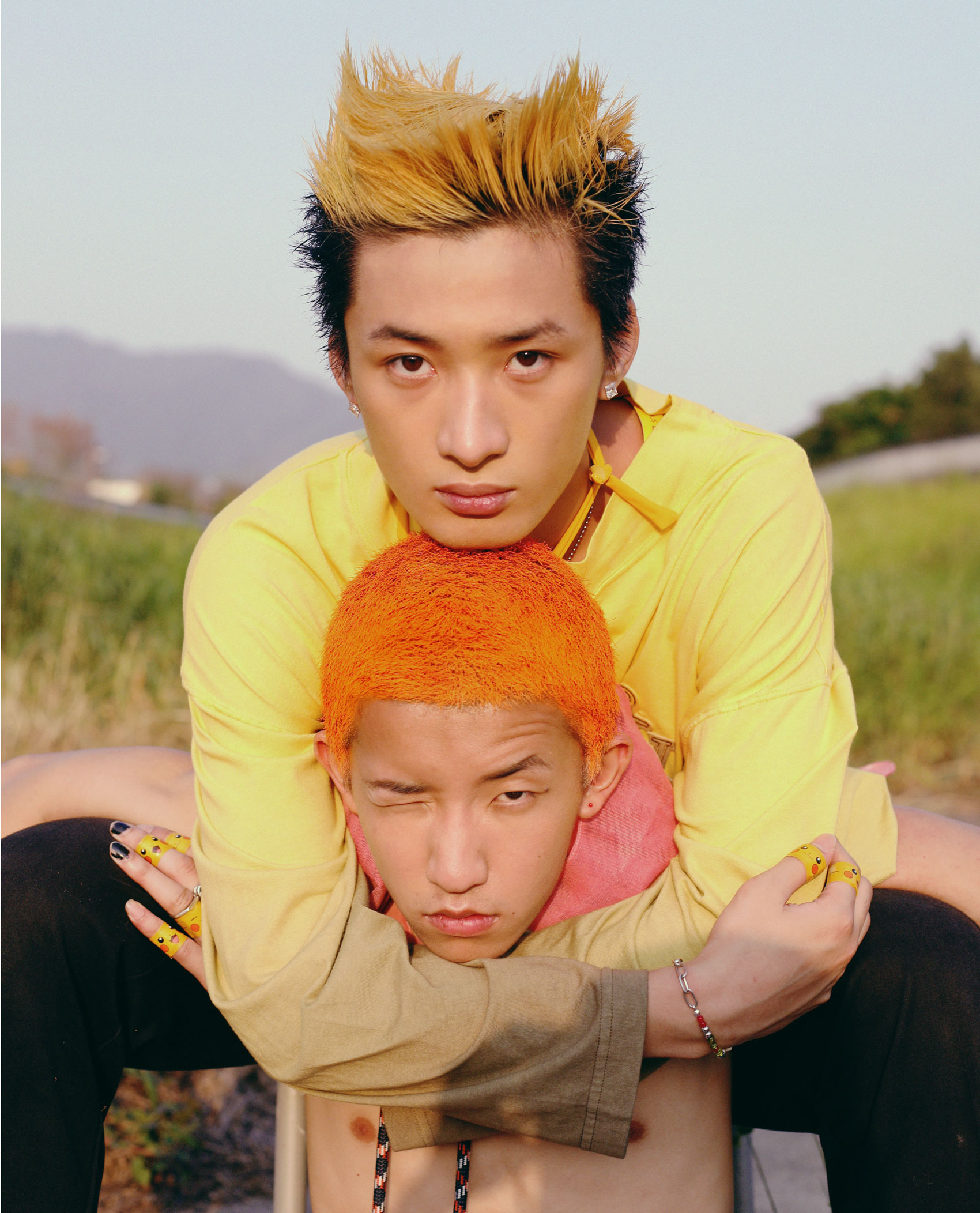 a young man with yellow hair sits behind a topless man with bright orange hair, wrapping his arms around him affectionately