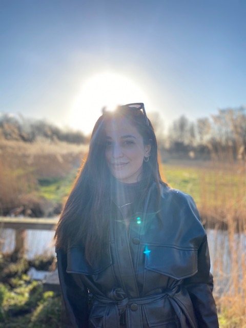 Noene – woman with long brown hair and a black leather coat, smiling and standing in front of the sun in a field.