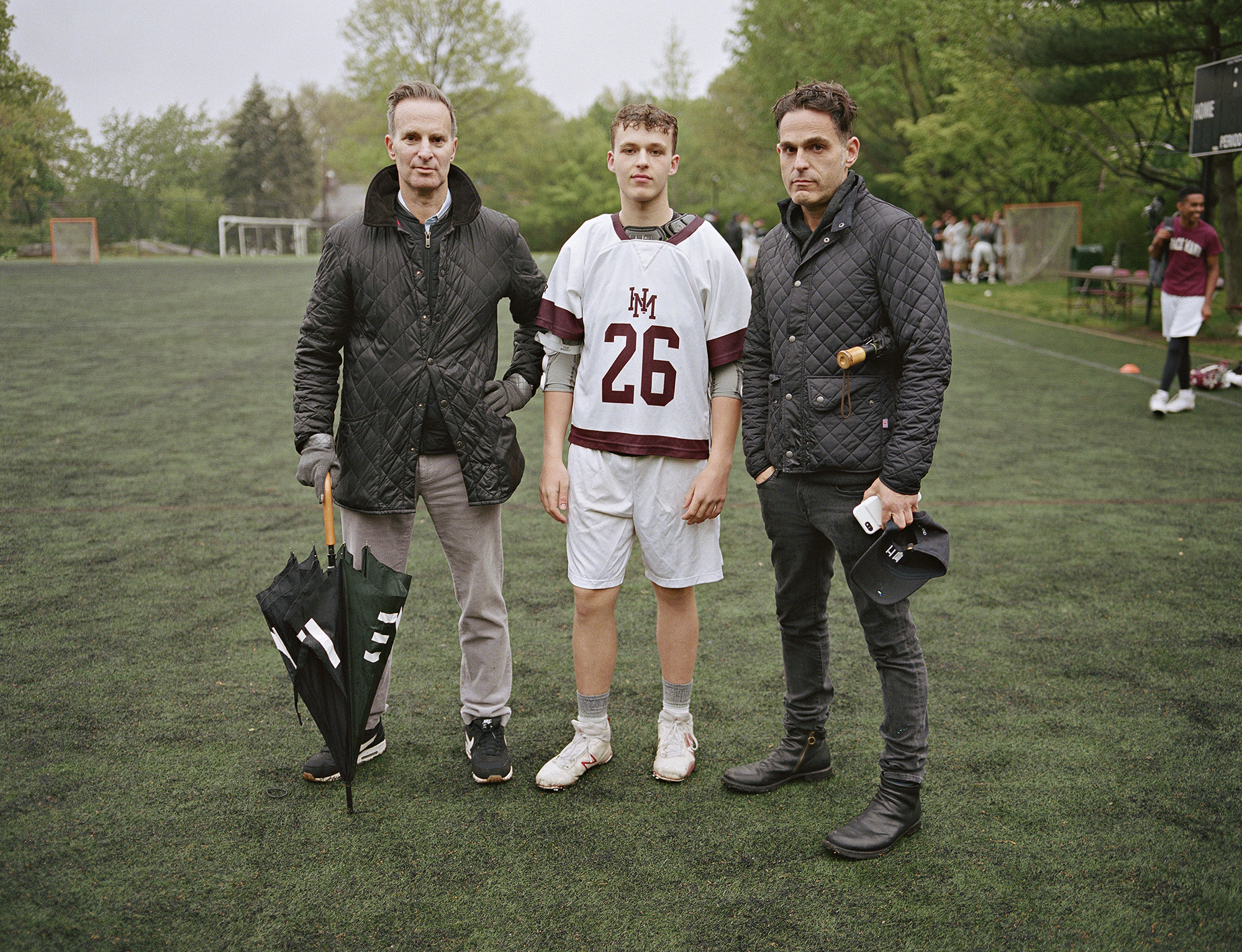 two men in quilted jackets stand on a football field with their son in a football jersey between them