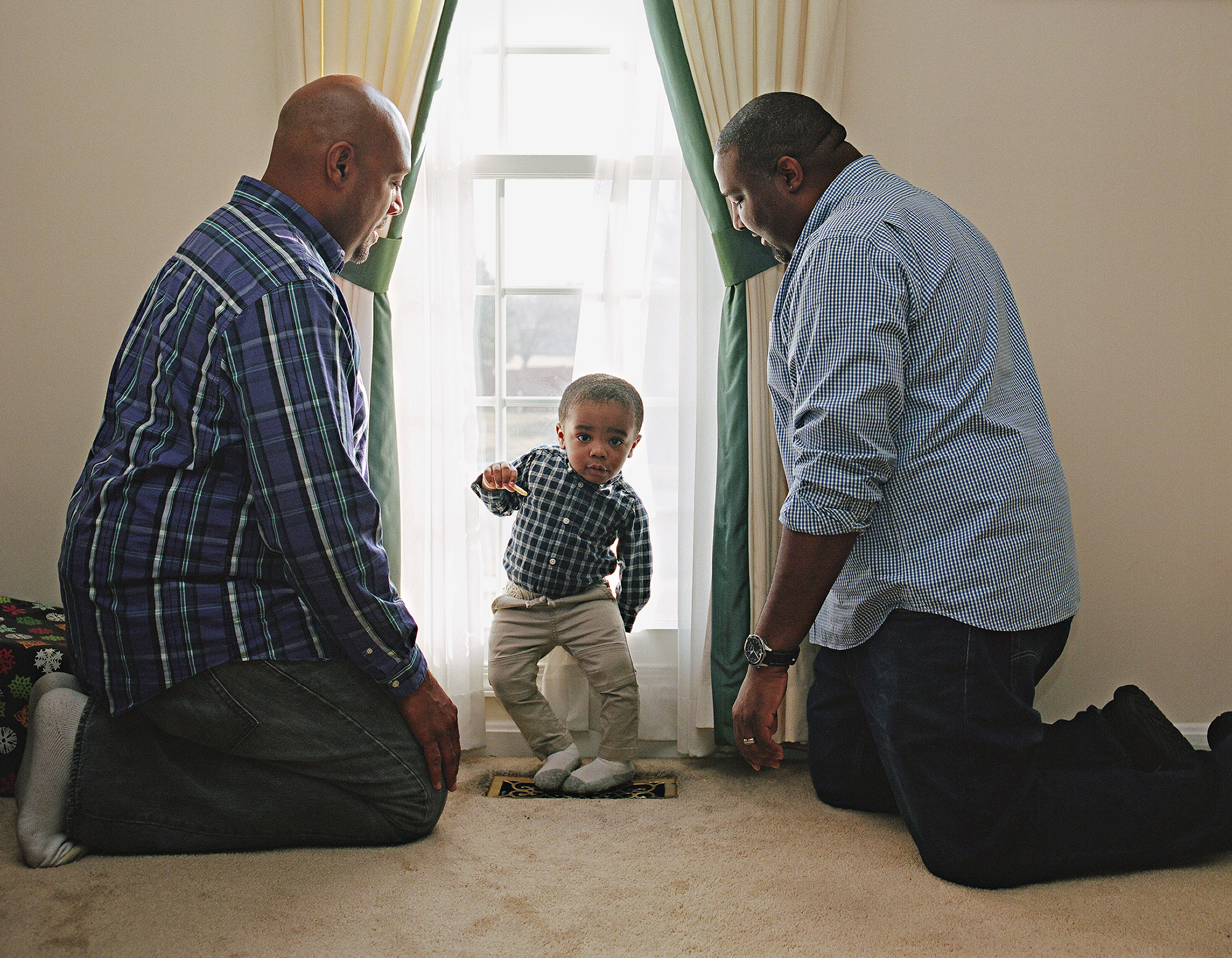 two men in patterned shirts look over their toddler son who stands in a window