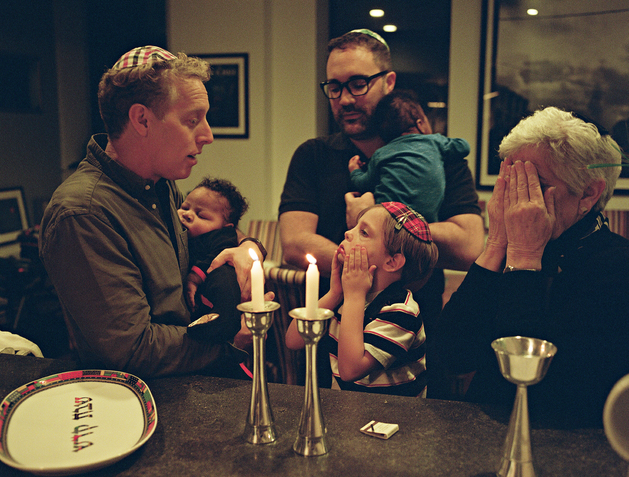 two fathers wearing kippahs play games with their young son and his grandmother. one carries a newborn baby.