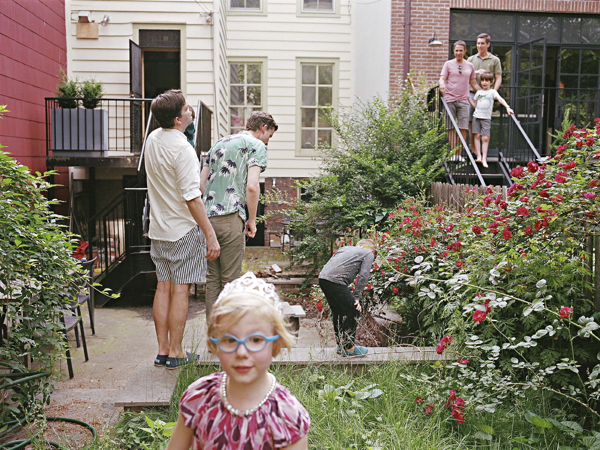 two families with gay male parents interact in their back garden as their kids play in the grass and bushes