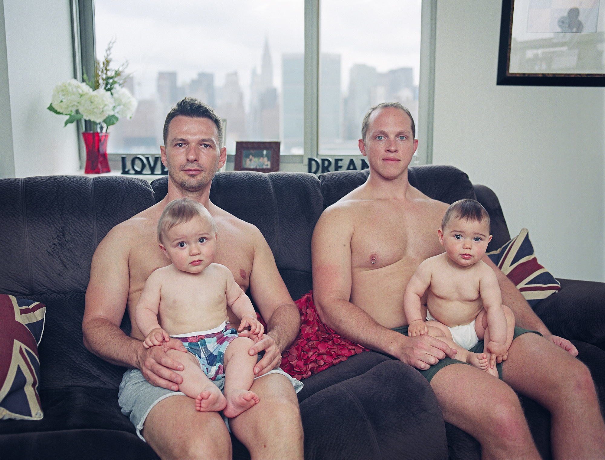 two gay fathers sit on their sofa with union jack cushions wearing no tops, each cradling one of their two children