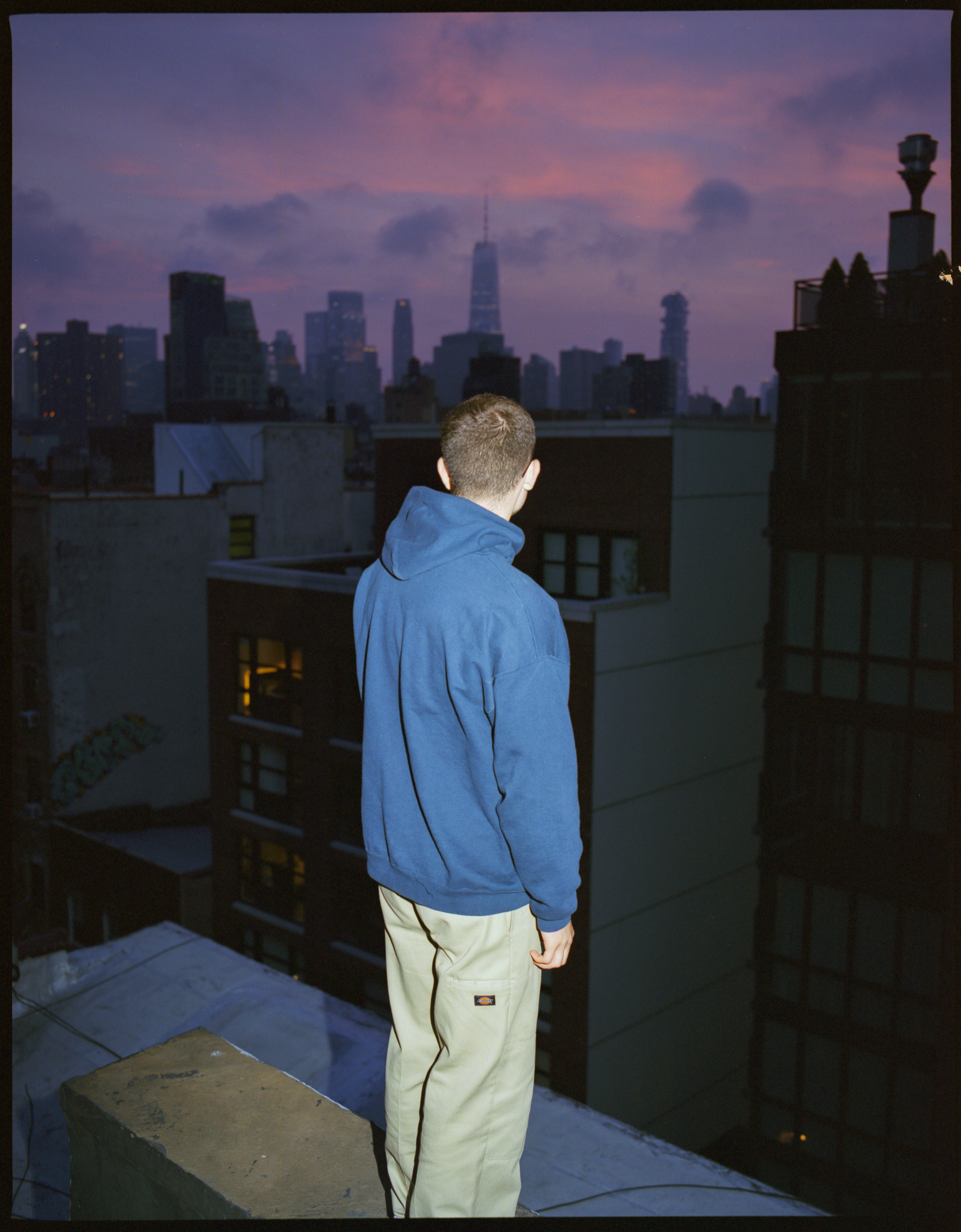 Man in blue hoodie standing on a roof over the purple New York sunset