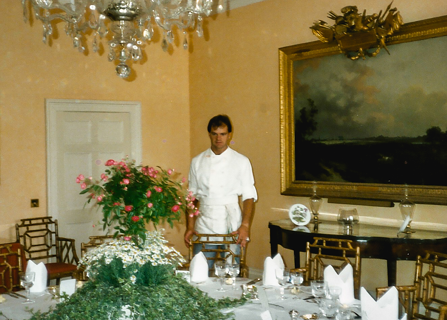 Enrico Derflingher – young man standing in an opulent dining room, wearing a white chef’s uniform and adjusting golden chairs around a large table.