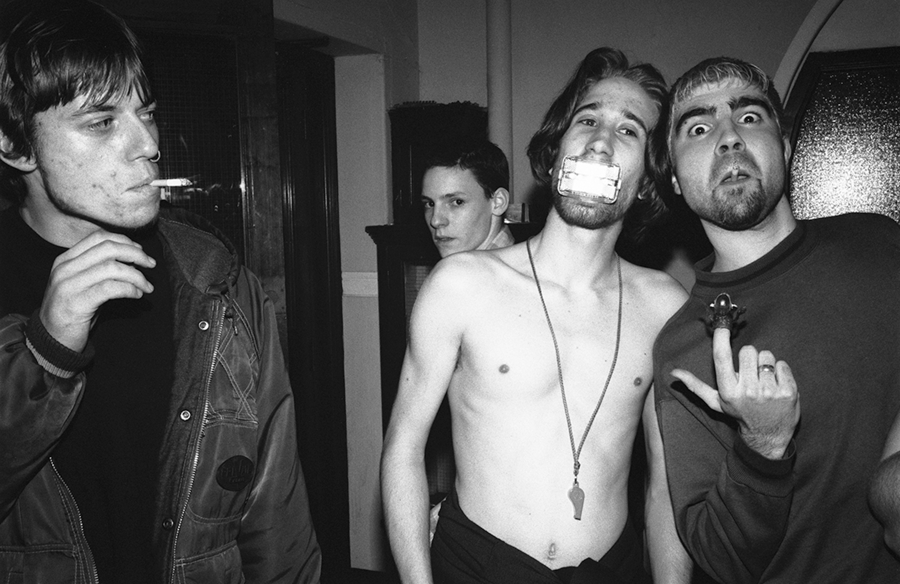 four men in a club, two smoking, one topless with a whistle around his neck and something in his mouth