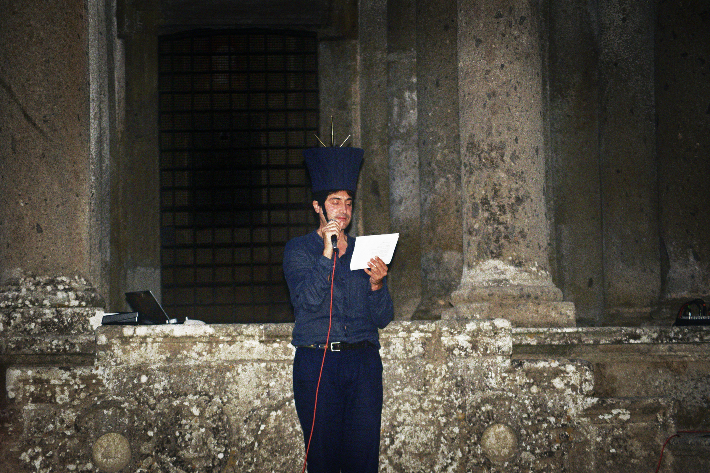 persona con in mano un microfono che legge qualcosa mente indossa uno strano cappello