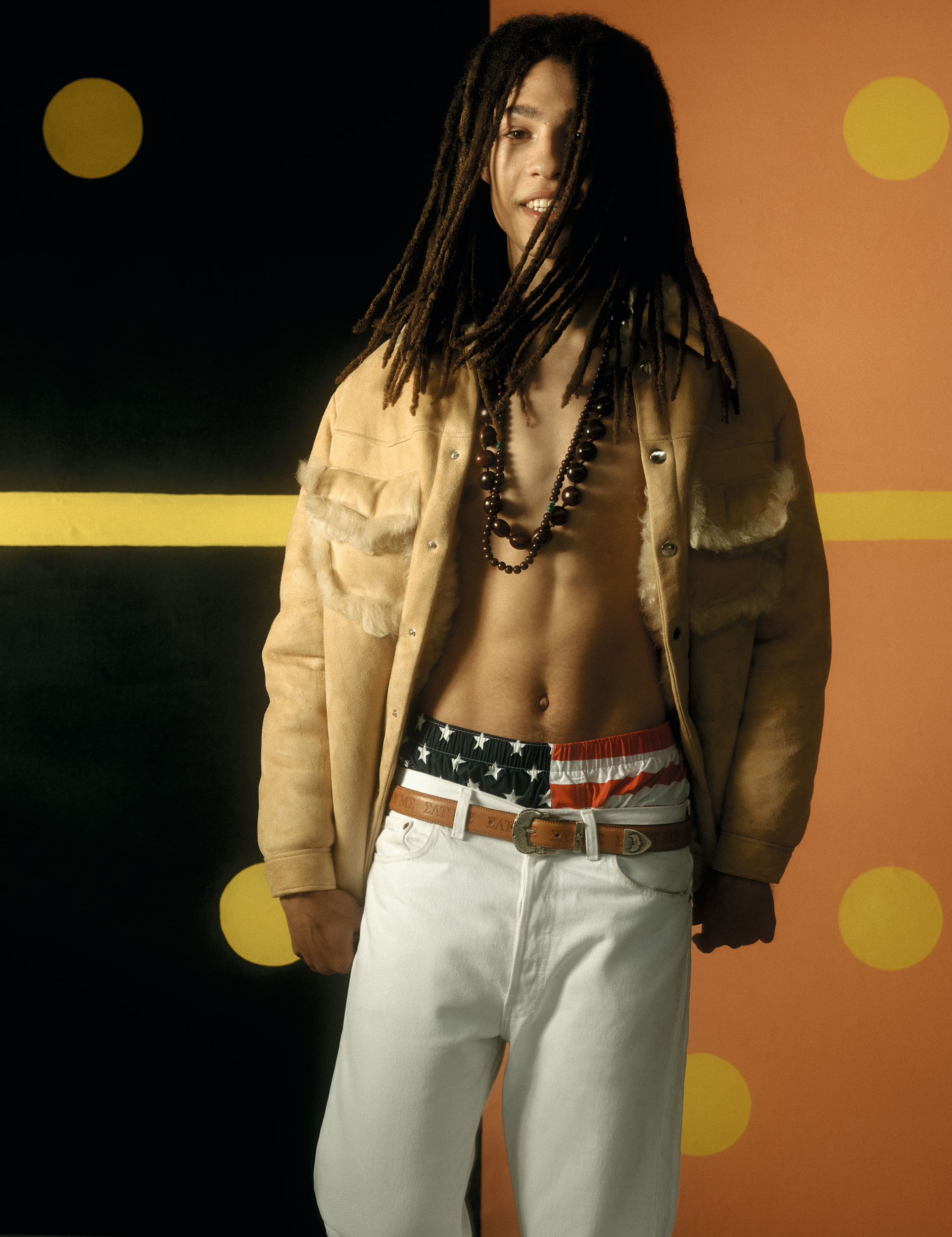 a model wearing white jeans with American flag boxers showing, with an open beige suede jacket and pearls 