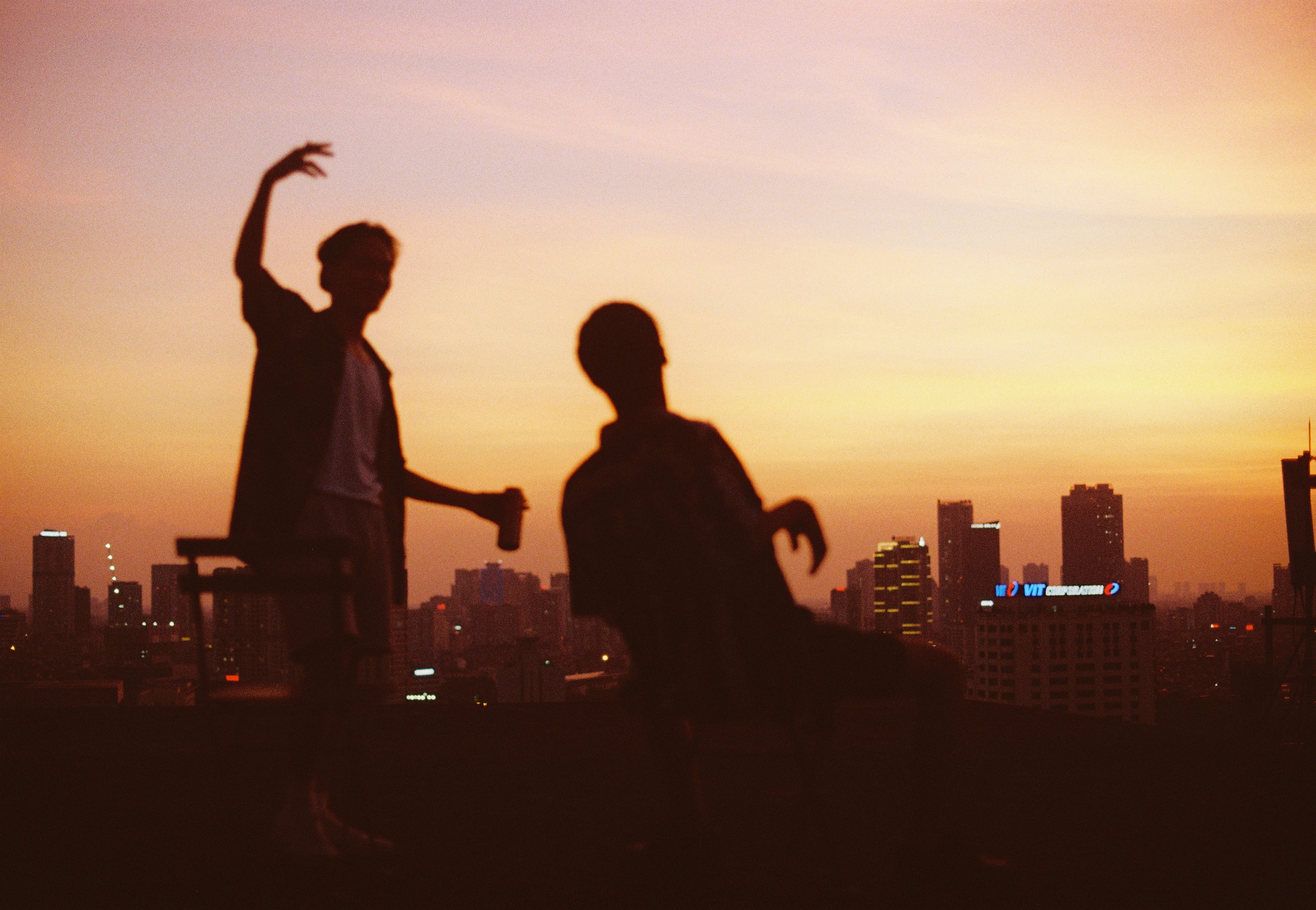 Two boys with cans of drinks dance around on a rooftop against the city skyline at sunset. 