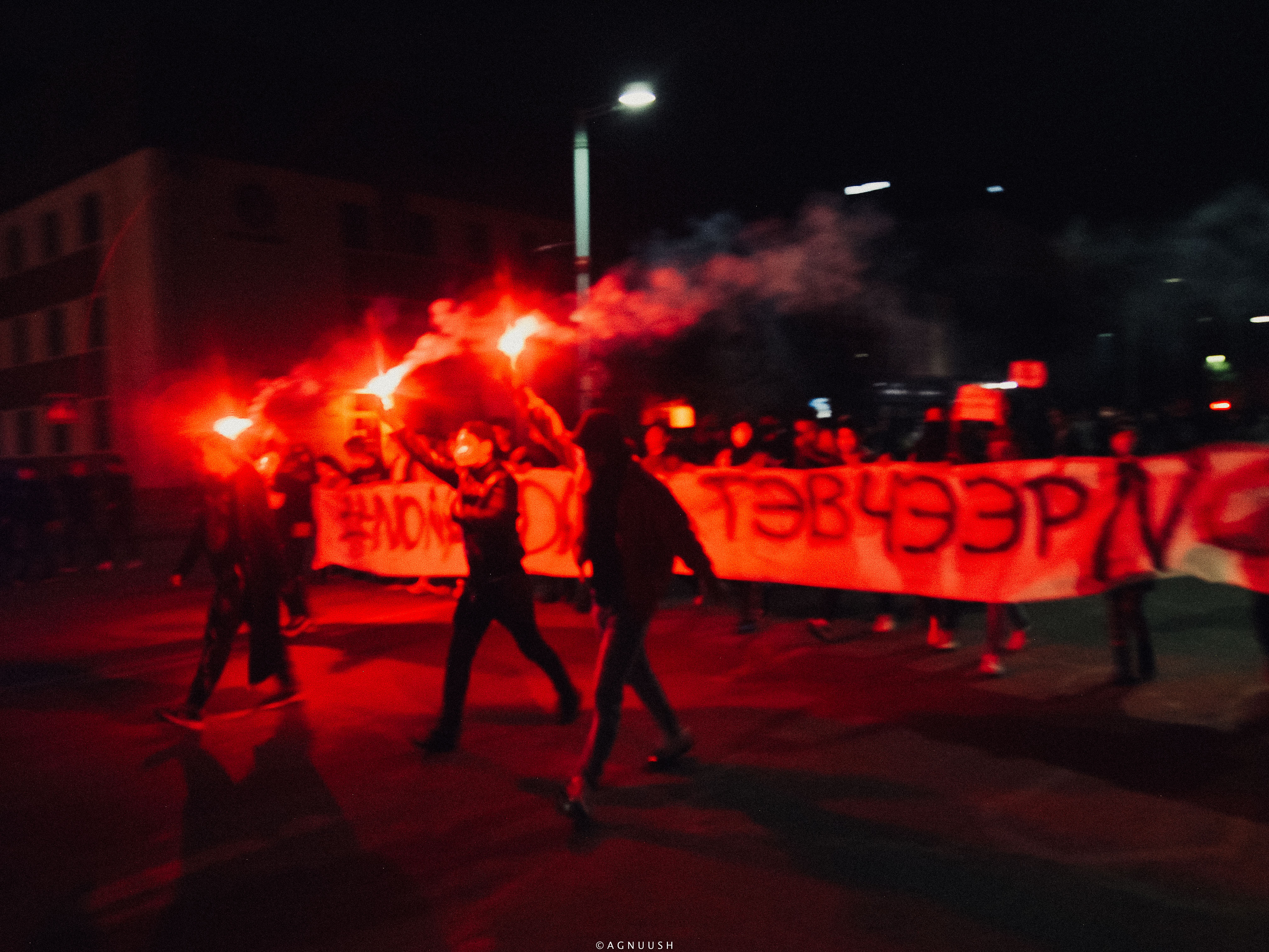 Large protest banner stretched across a road and lit under red flares.