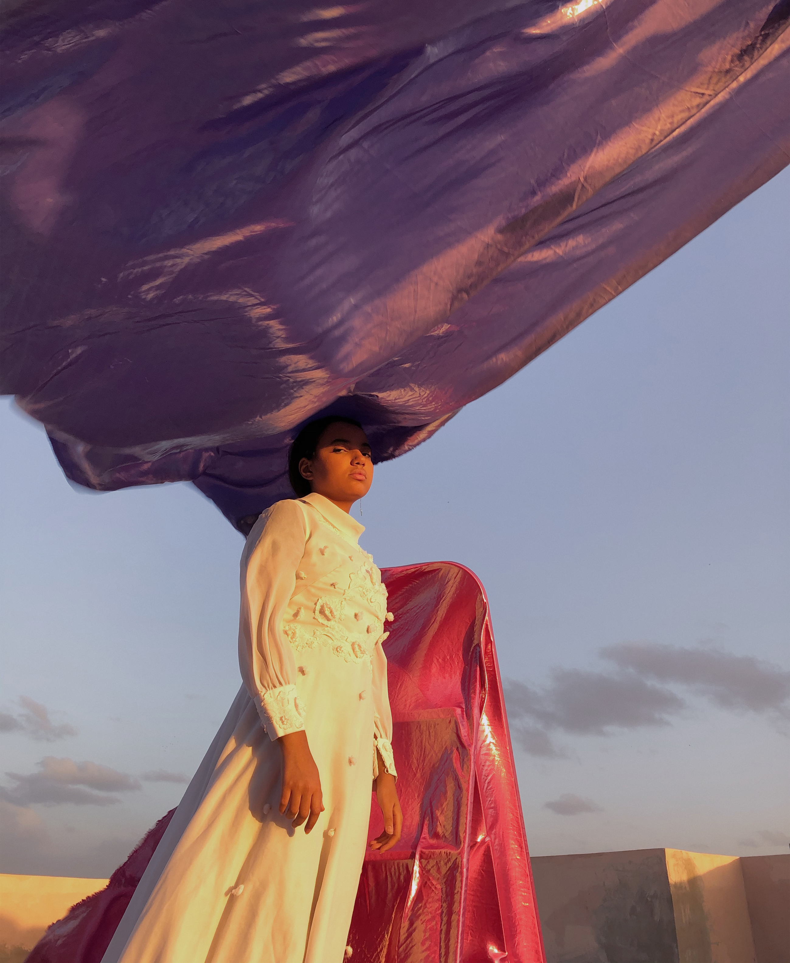 An individual in a white sleeved gown against the setting sky with neon metallic pink and purple inflatables behind them and resting on their head.