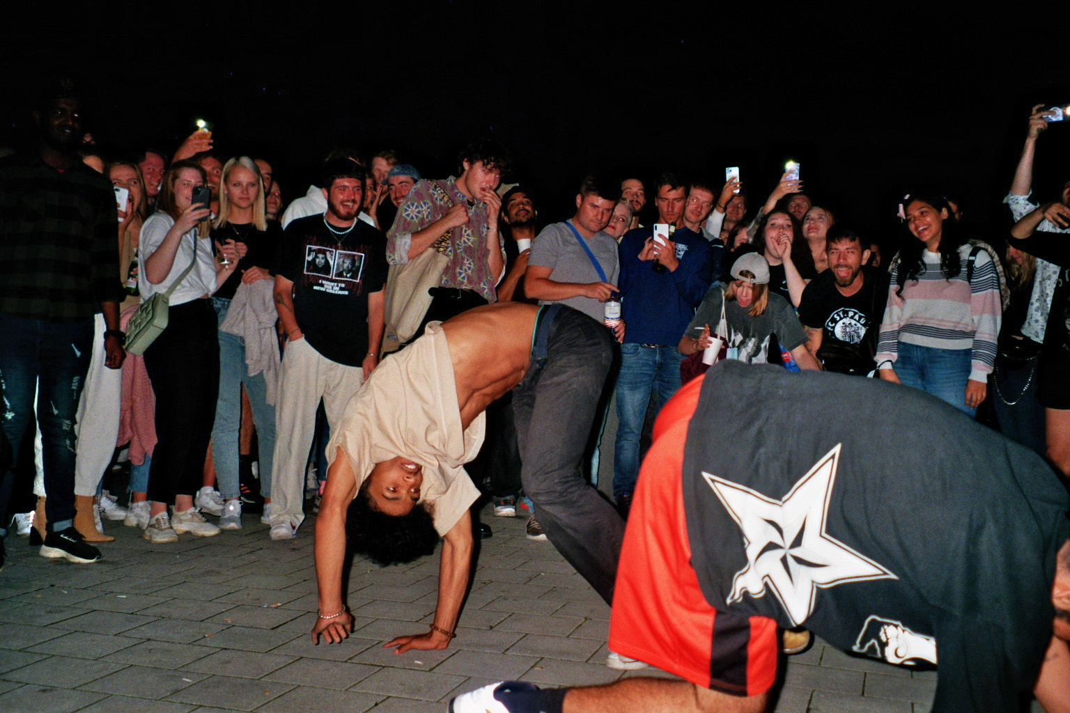 A crowd filming two people doing handstands and dancing on the pavement. 