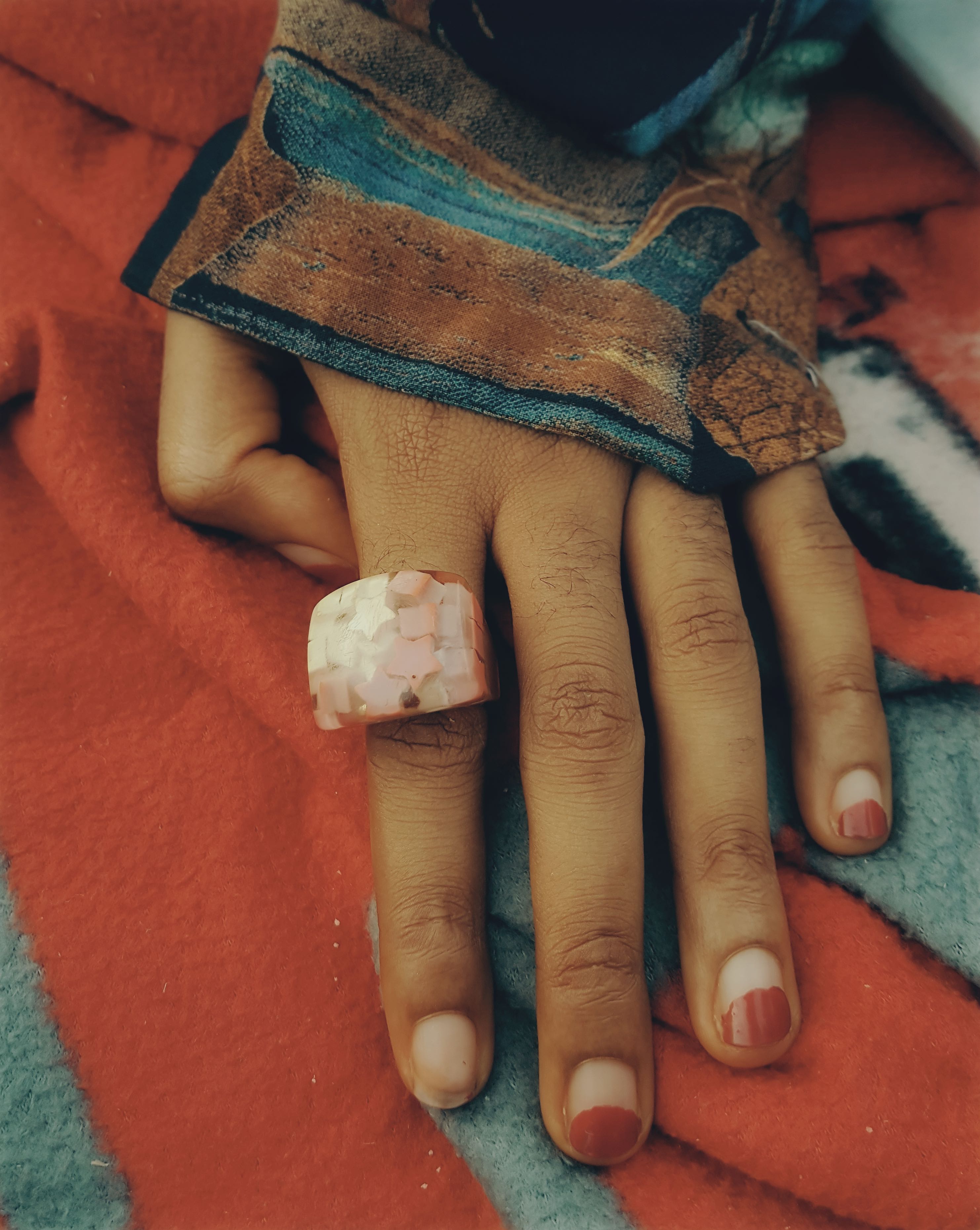 A hand on a patterned sheet with half painted nails and a large pink stone ring. 
