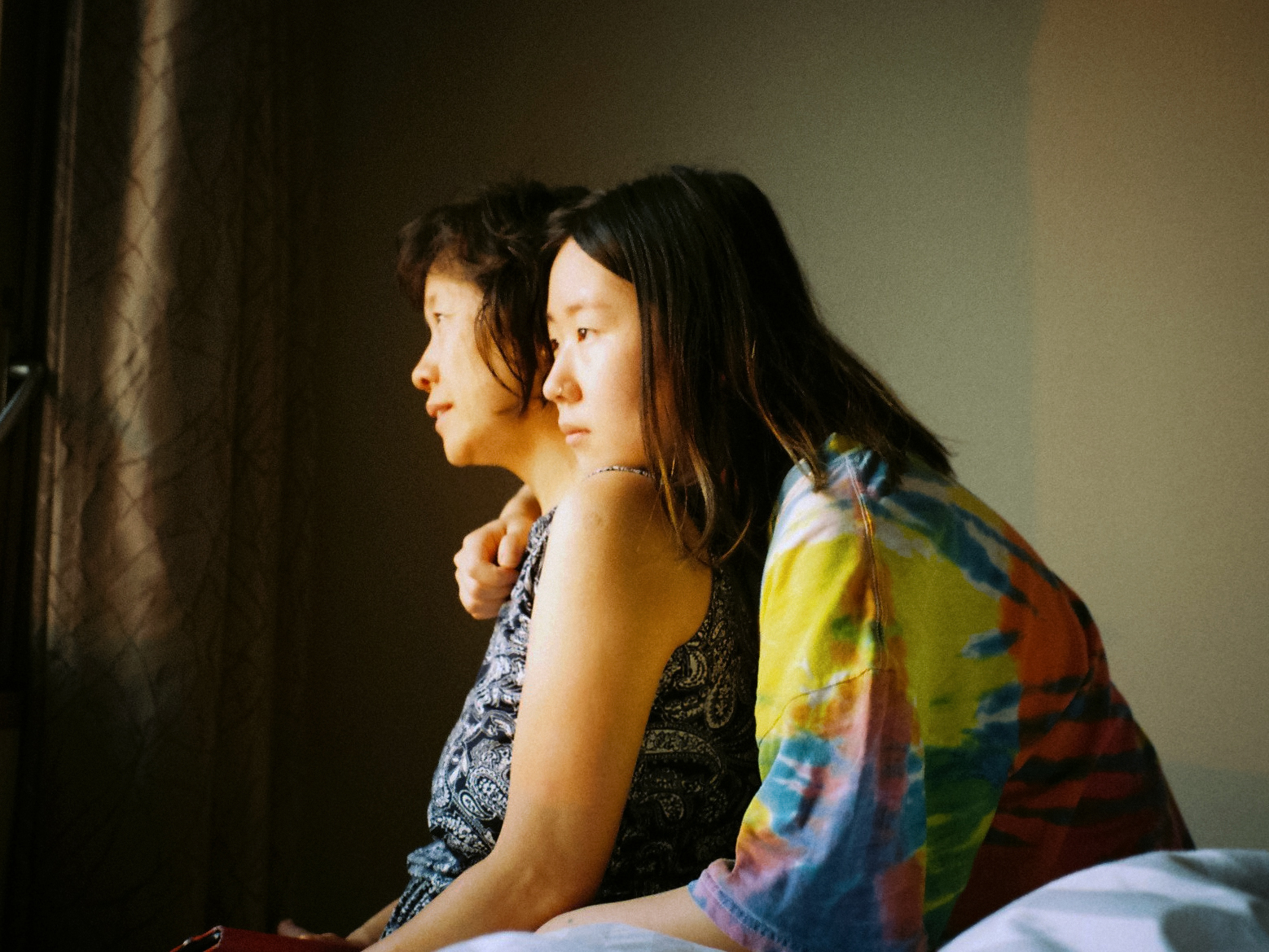a young girl and her mum sit and look out the window. She wear a tie-dye top and her mum wears a black patterned blouse. 